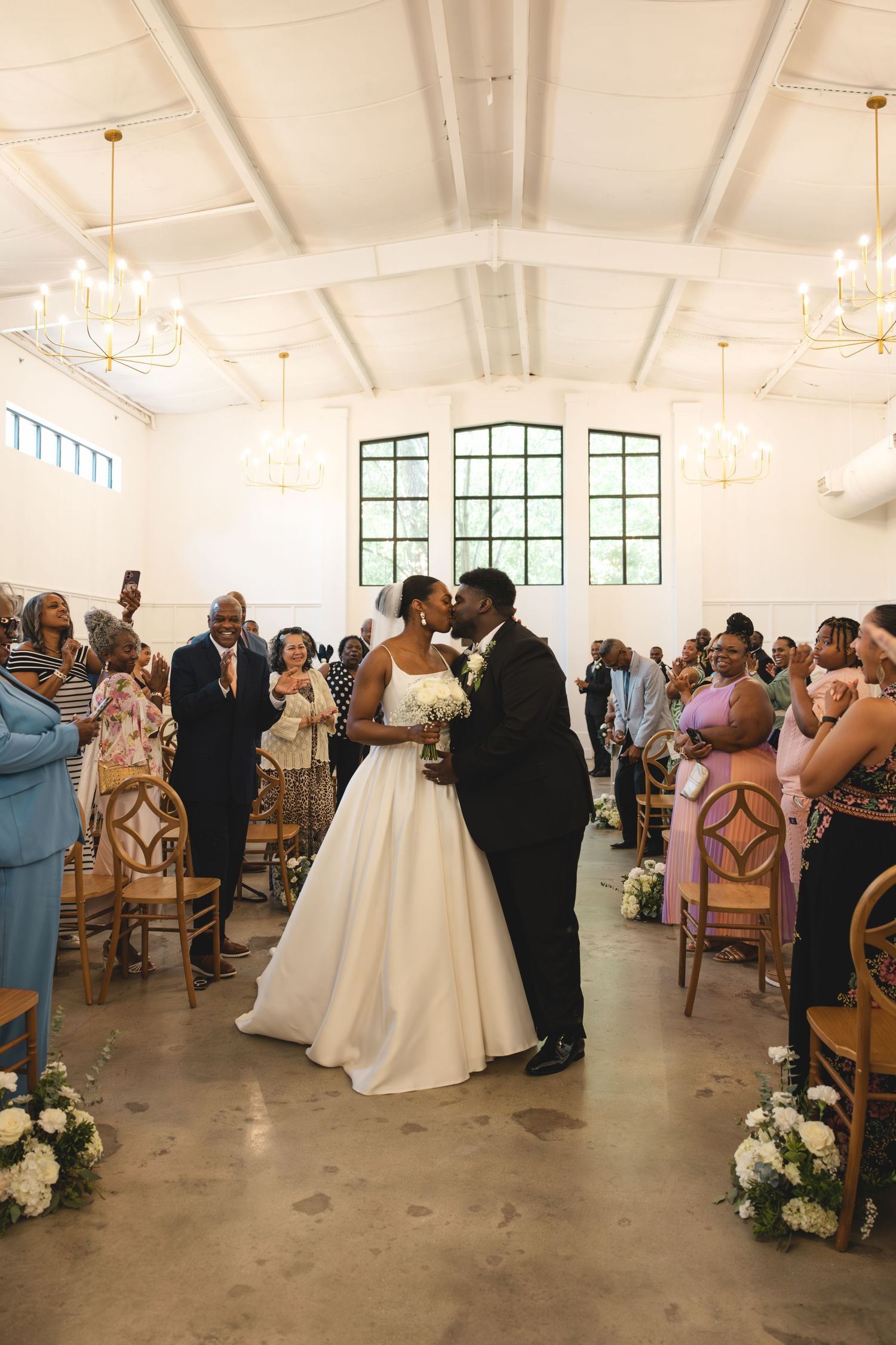 Newlyweds share first kiss in aisle of bright white wedding venue surrounded by cheering guests.