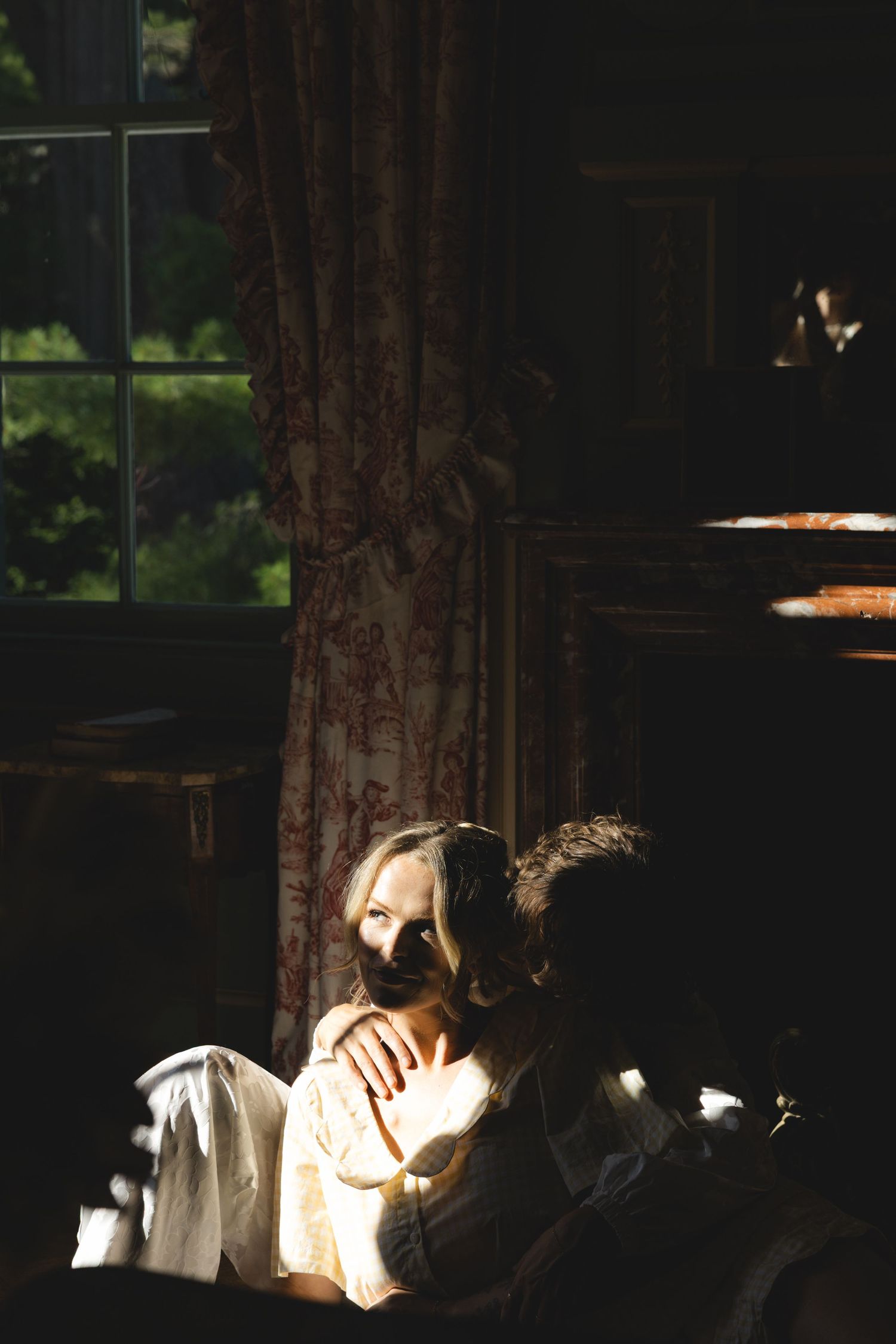 A couple embraces in dramatic chiaroscuro lighting inside a dark vintage room with floral curtains and window.