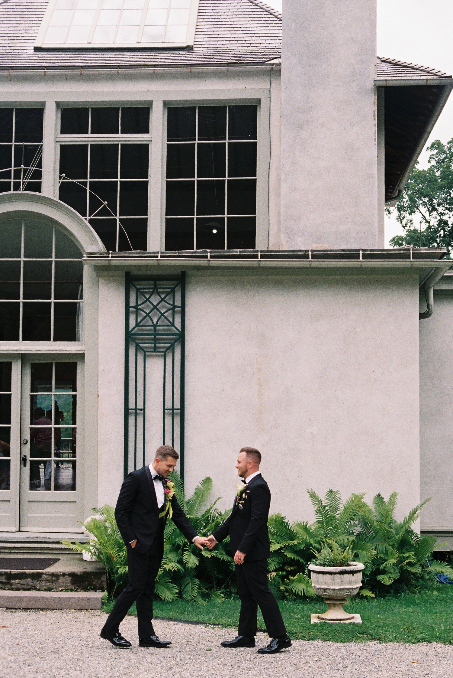 Two grooms in black tuxedos share a joyful moment outside an elegant estate during their wedding day.