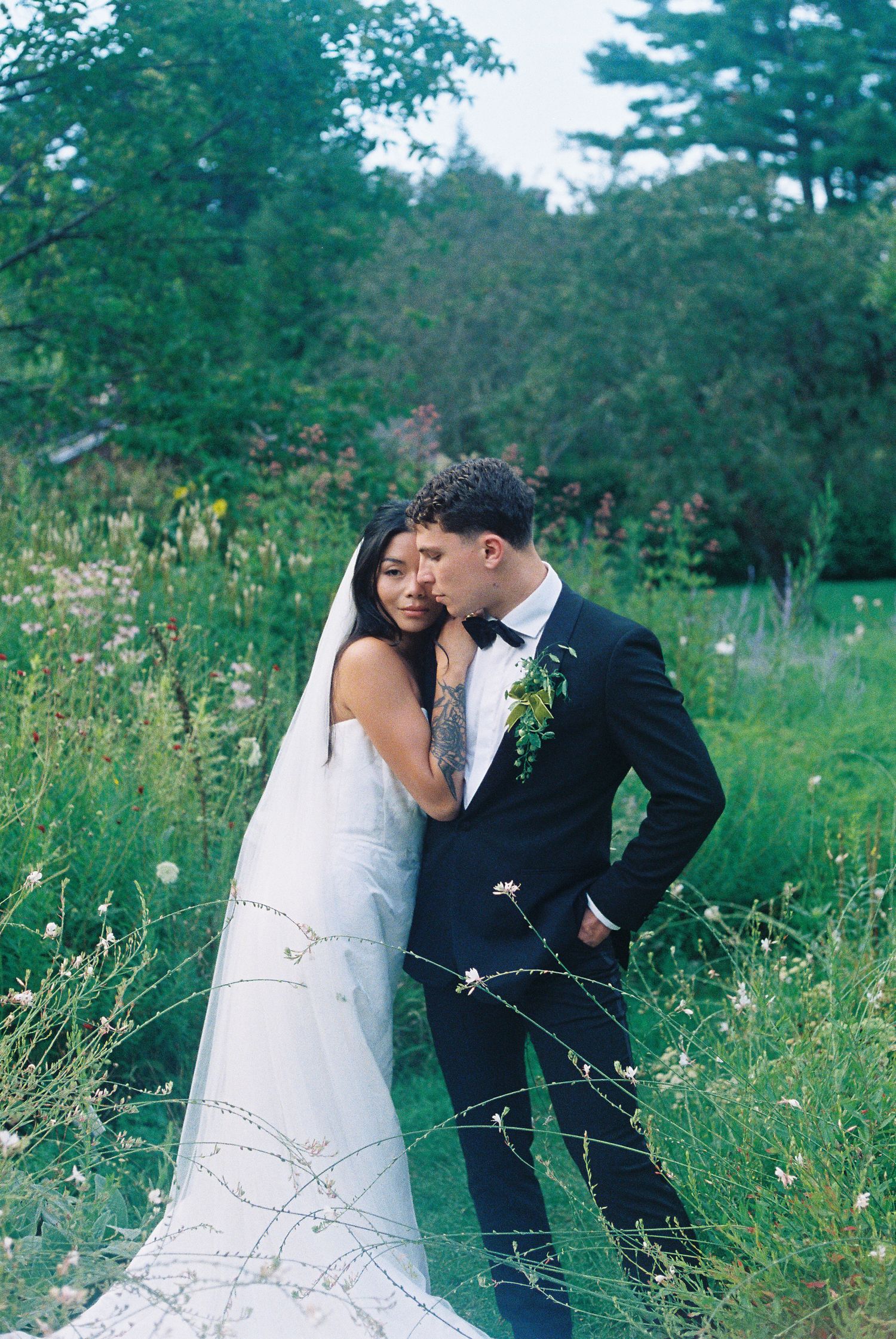 Bride and groom embrace in a lush garden, wearing a white gown with veil and black tuxedo with boutonniere.