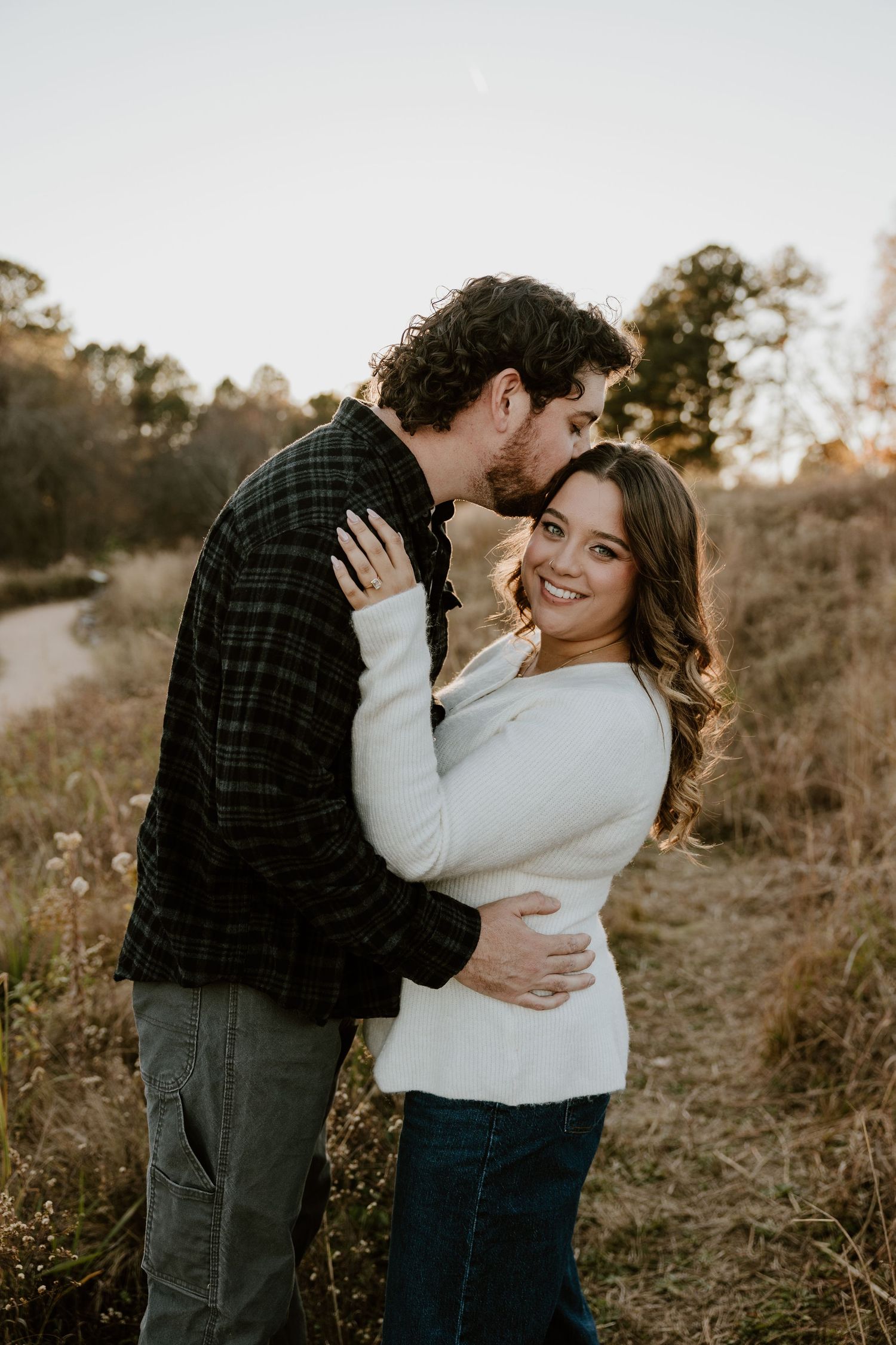 Couple embracing in a golden field at sunset, woman in white sweater smiling, man in plaid shirt kissing her cheek.