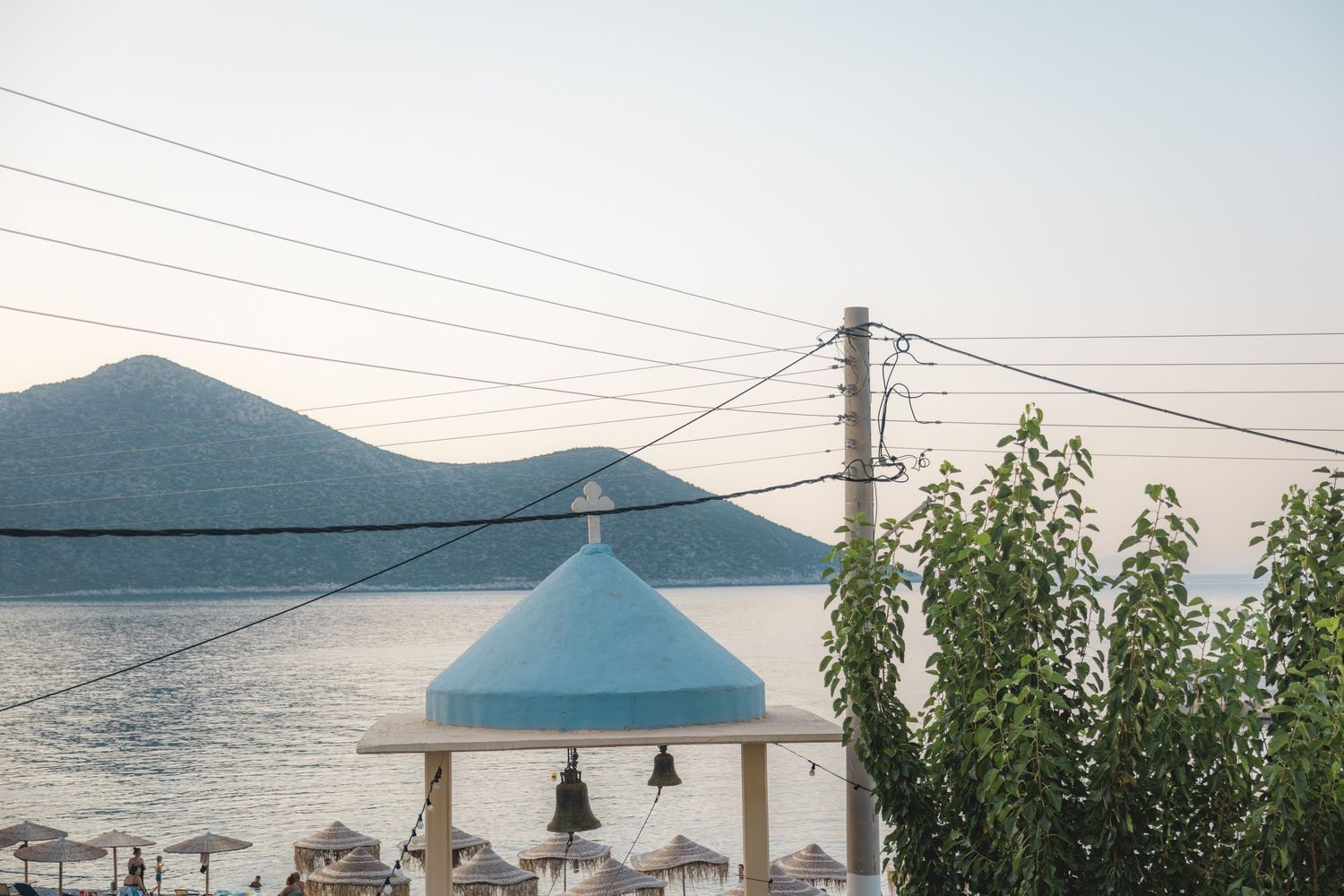Blue-domed chapel near a Mediterranean seaside with mountains, power lines, and beach umbrellas at sunset.