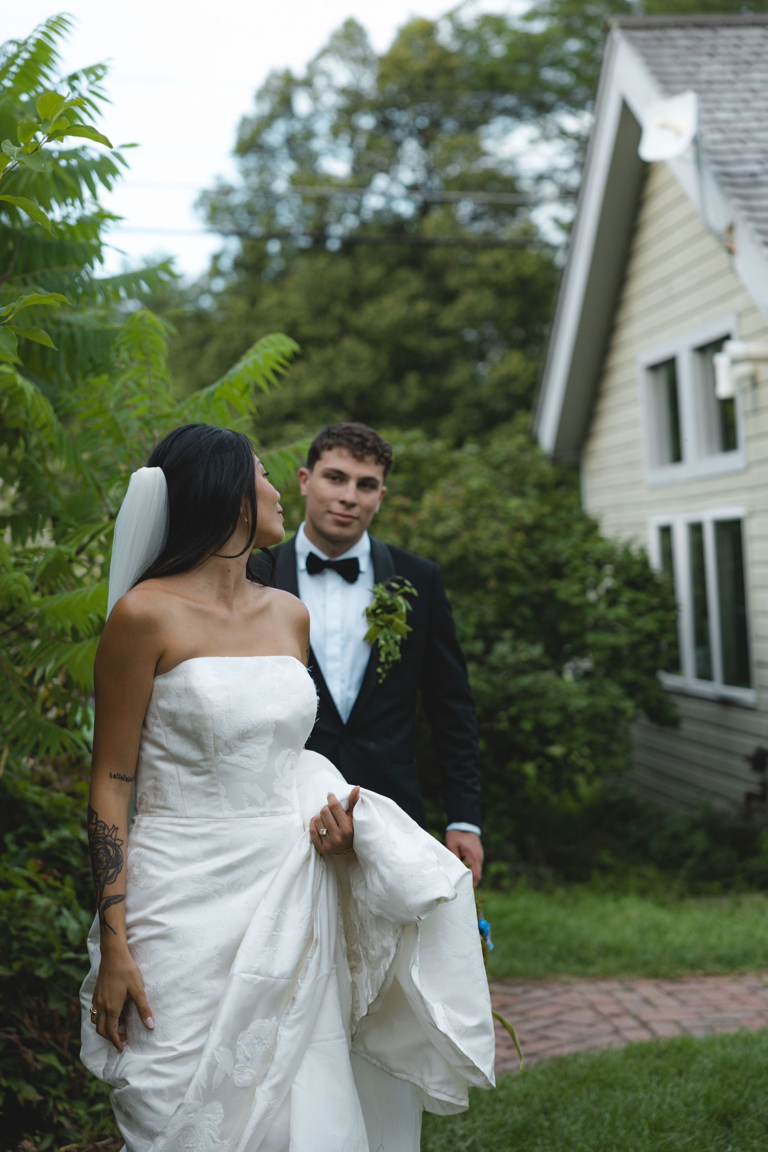 Bride in strapless white gown and groom in black tuxedo walk hand in hand outside a charming house surrounded by greenery.