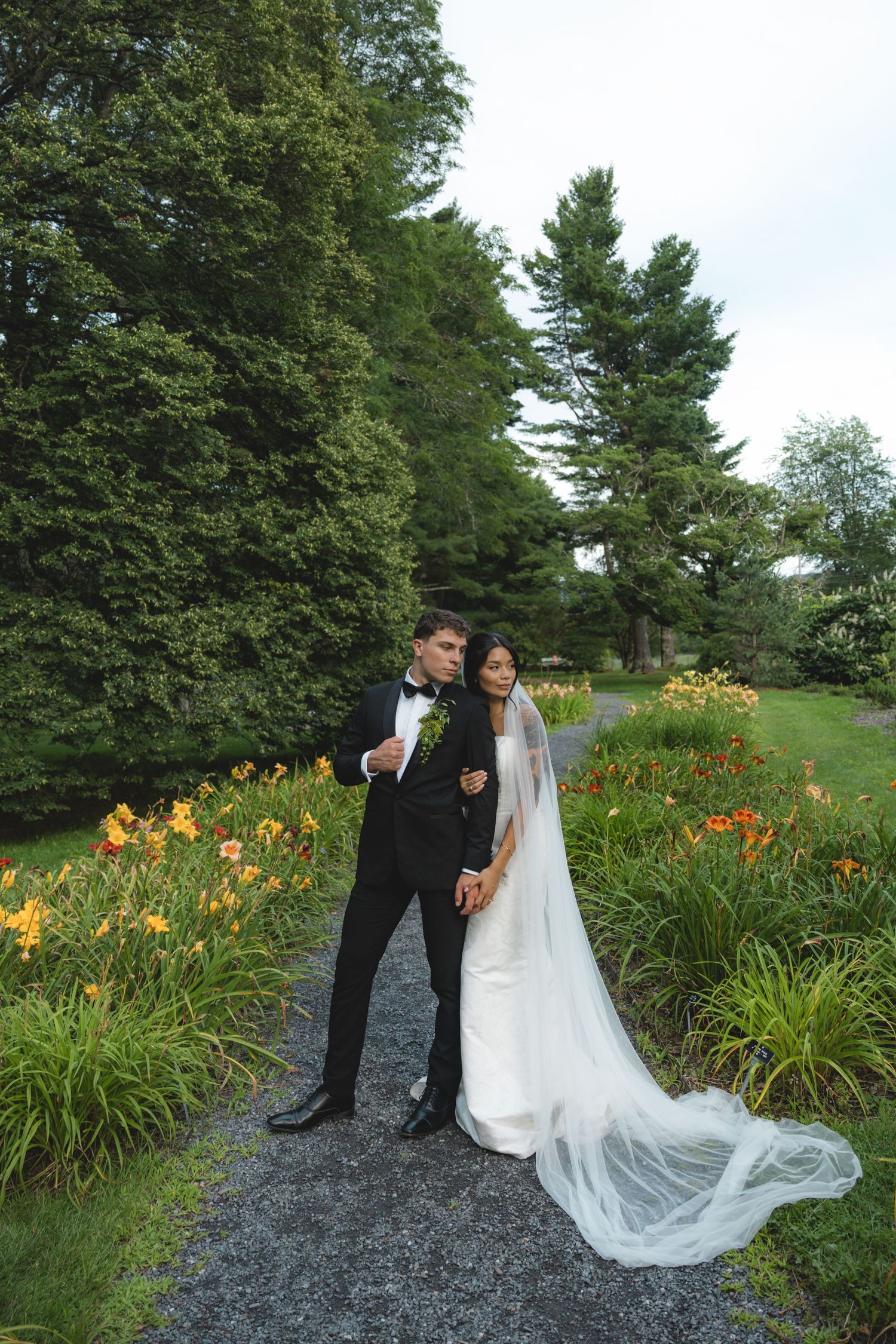 Bride and groom pose on garden path surrounded by colorful flowers and lush green trees on their wedding day.