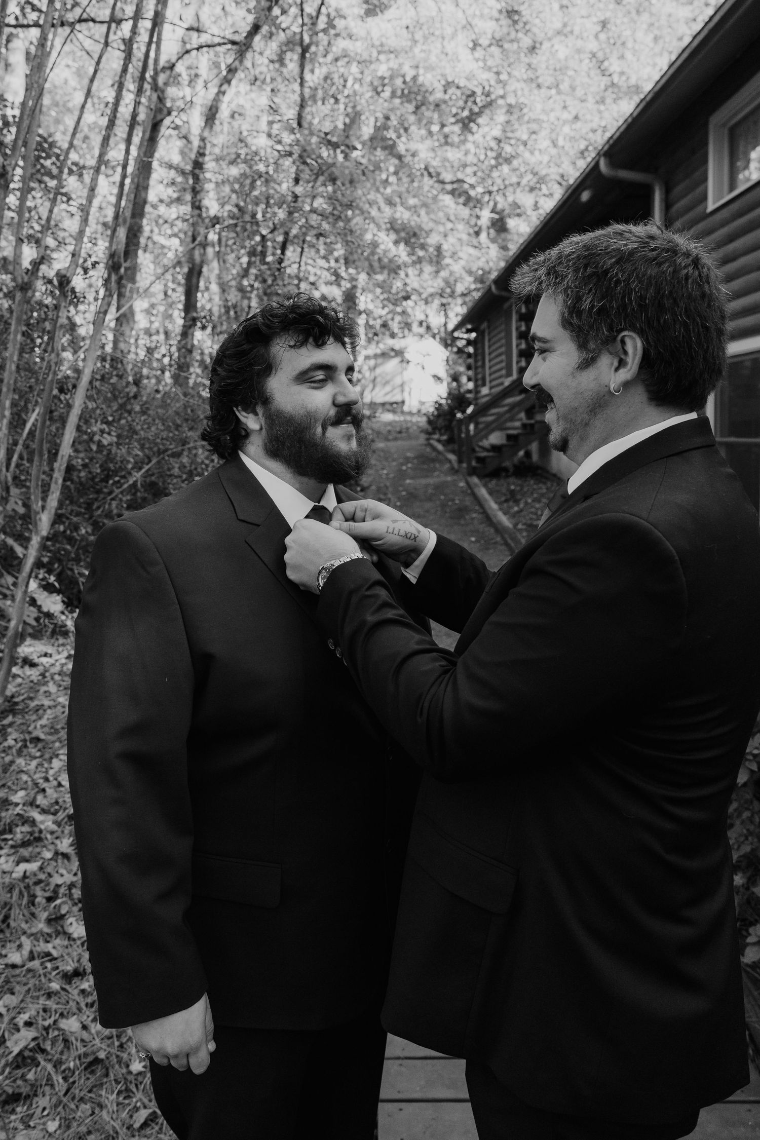 Black and white photo of a man adjusting another mans tie outdoors near a log cabin, likely at a wedding.