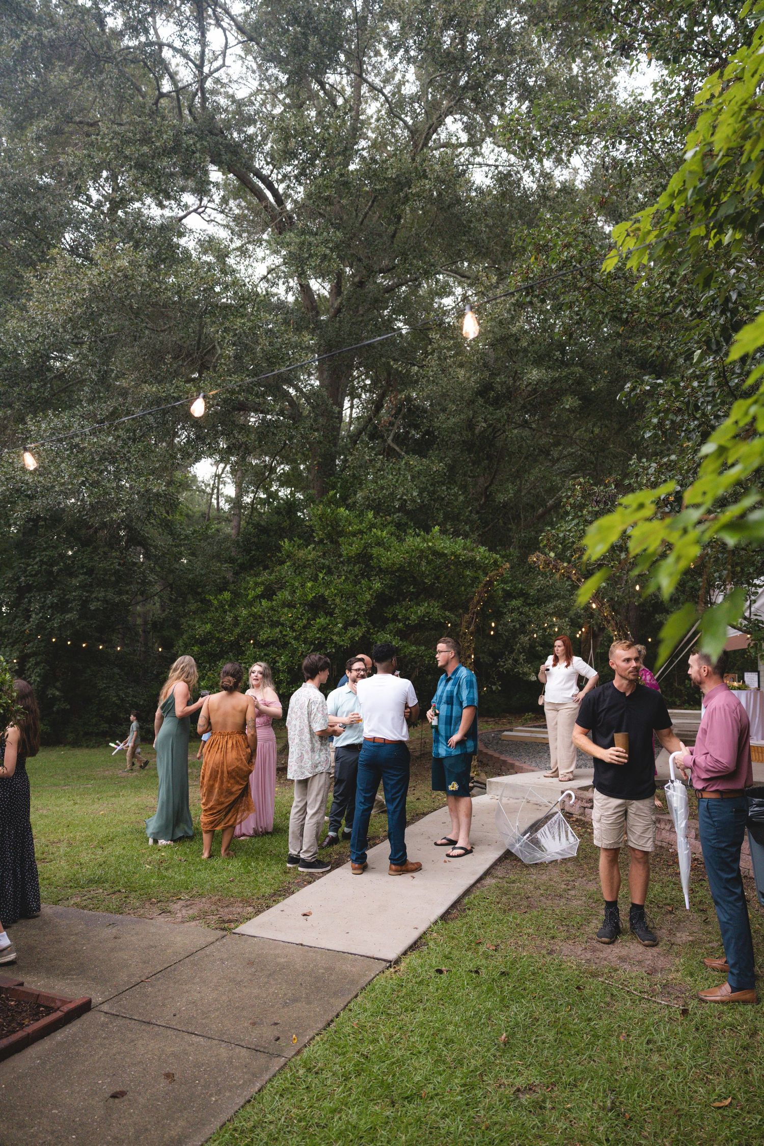 Guests mingle outdoors at a garden party with string lights and lush trees in the background.