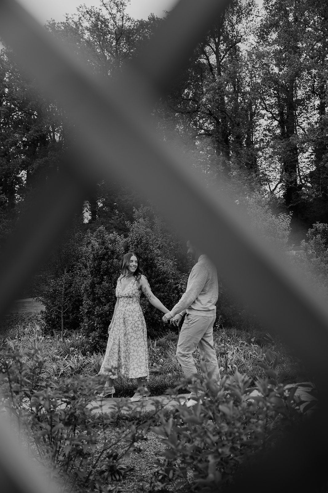 Black and white photo of a couple holding hands in a lush garden, woman in floral dress smiling at partner.