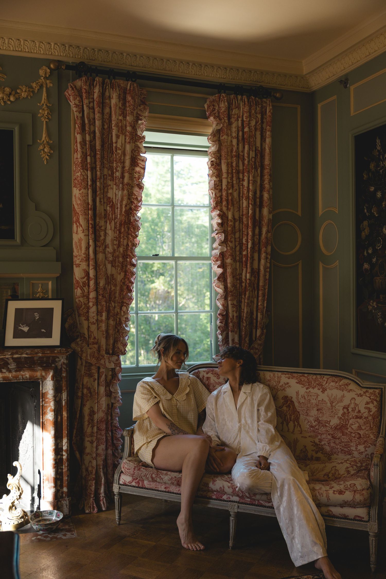 Two women in robes sit on a floral sofa by a tall window in an elegant, green-paneled room with ornate curtains.