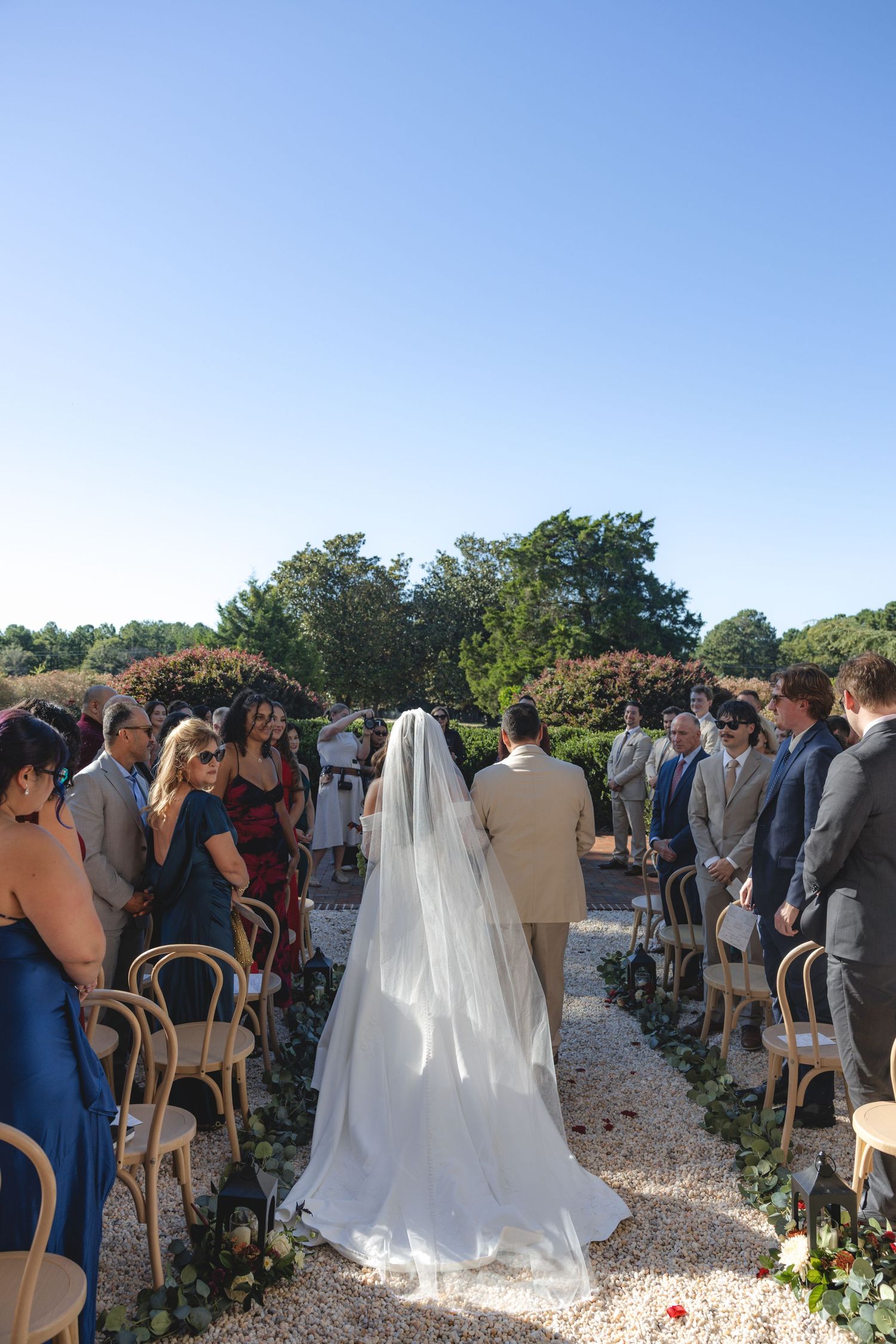 Bride walking down outdoor wedding aisle in white gown and veil, flanked by seated guests on a sunny day.