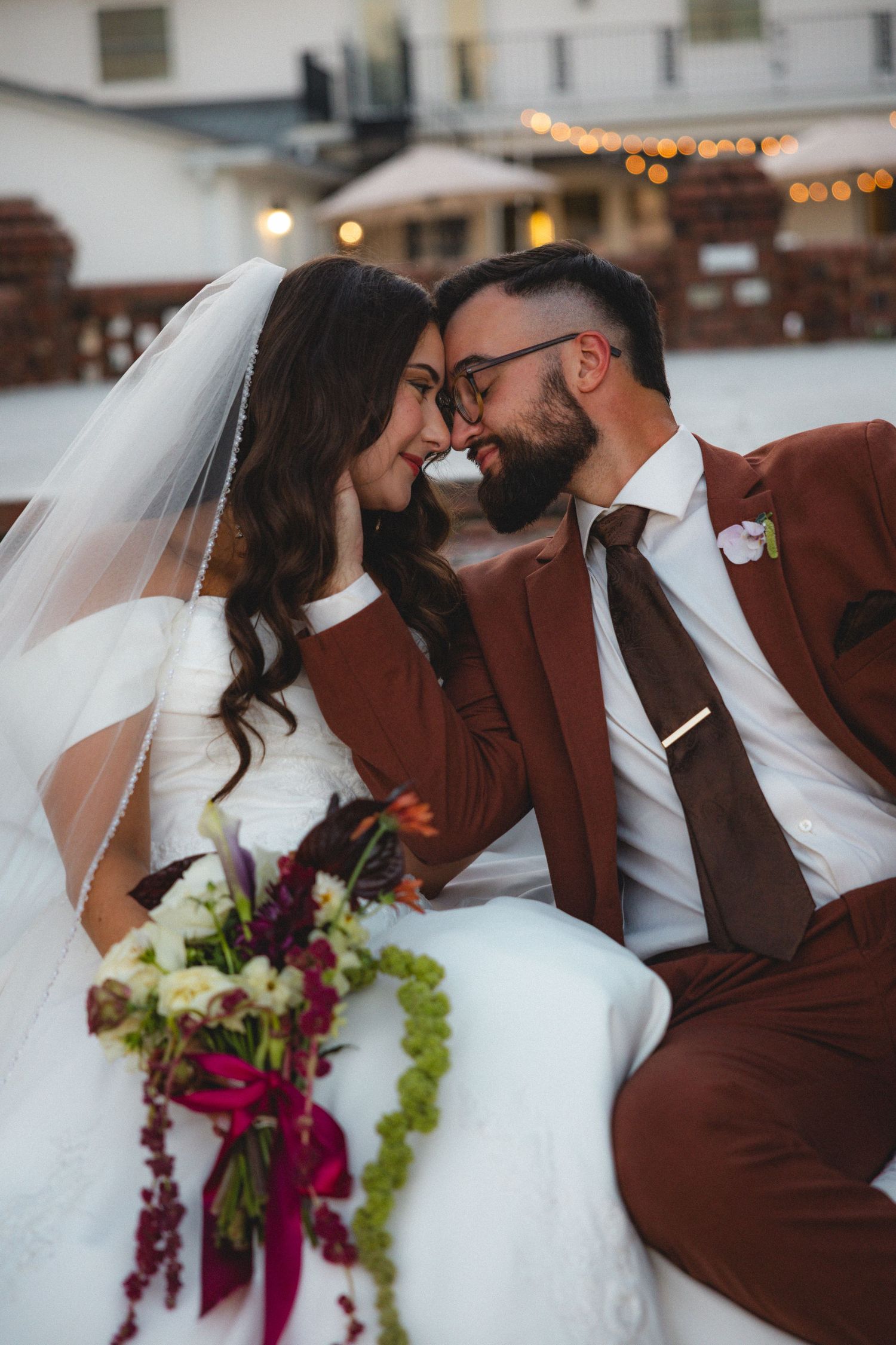 A bride and groom share a romantic nose-to-nose moment, she in a white gown with veil, he in a brown suit.