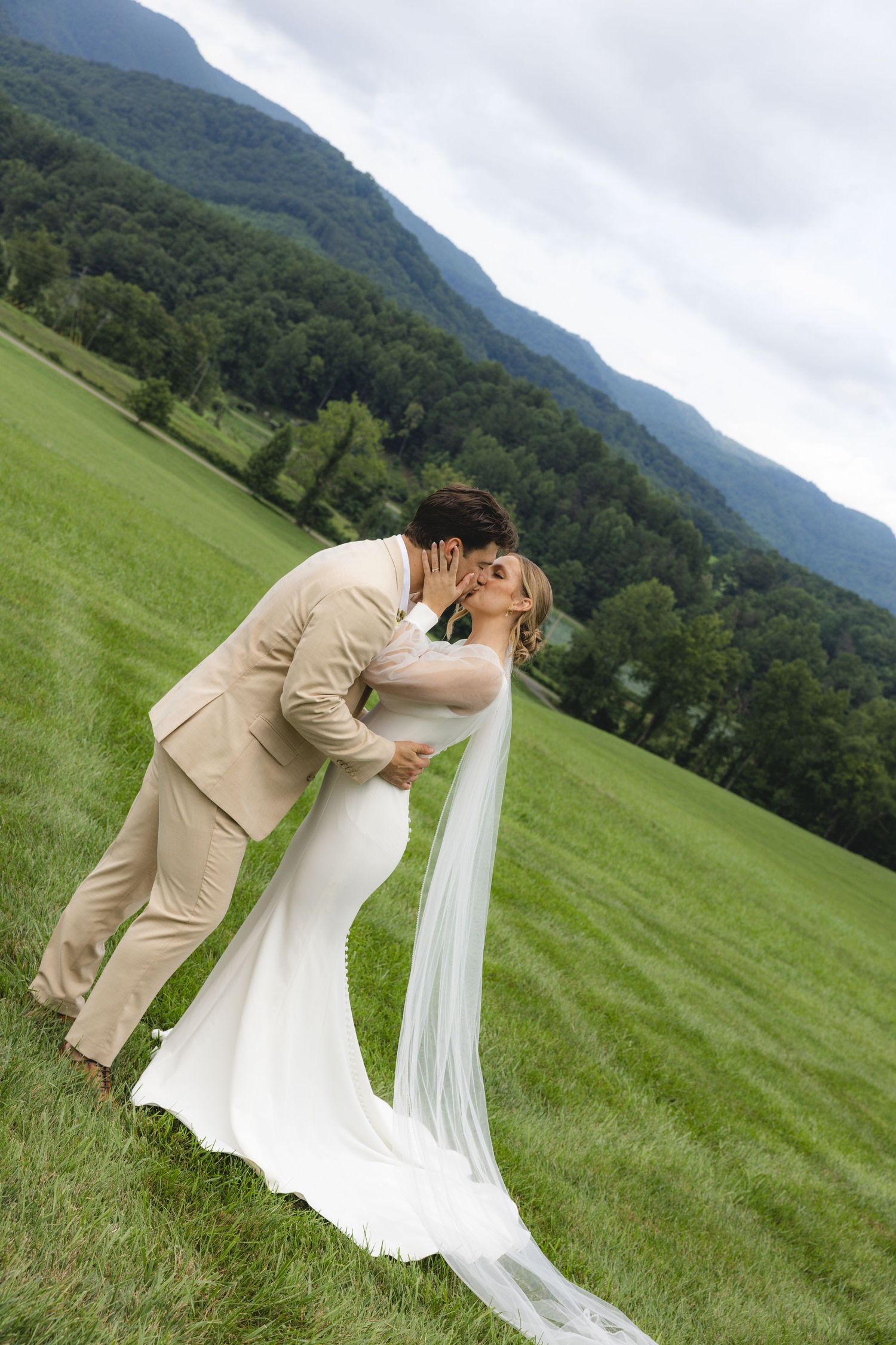 A bride and groom share a romantic dip kiss in a lush green meadow with misty mountains in the background.
