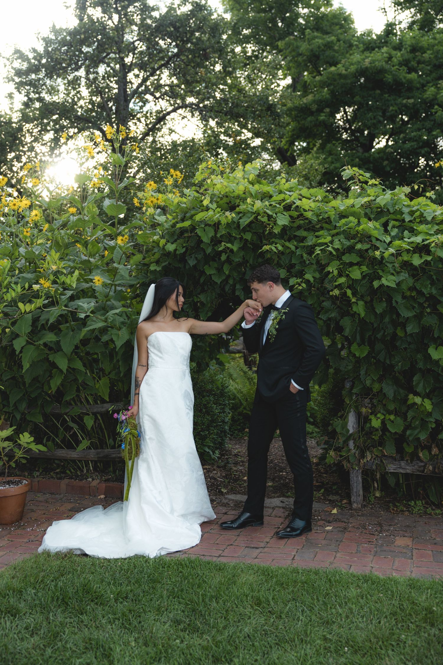 Groom in black tuxedo kisses bride's hand in lush garden setting, bride wearing white gown with veil holding bouquet.