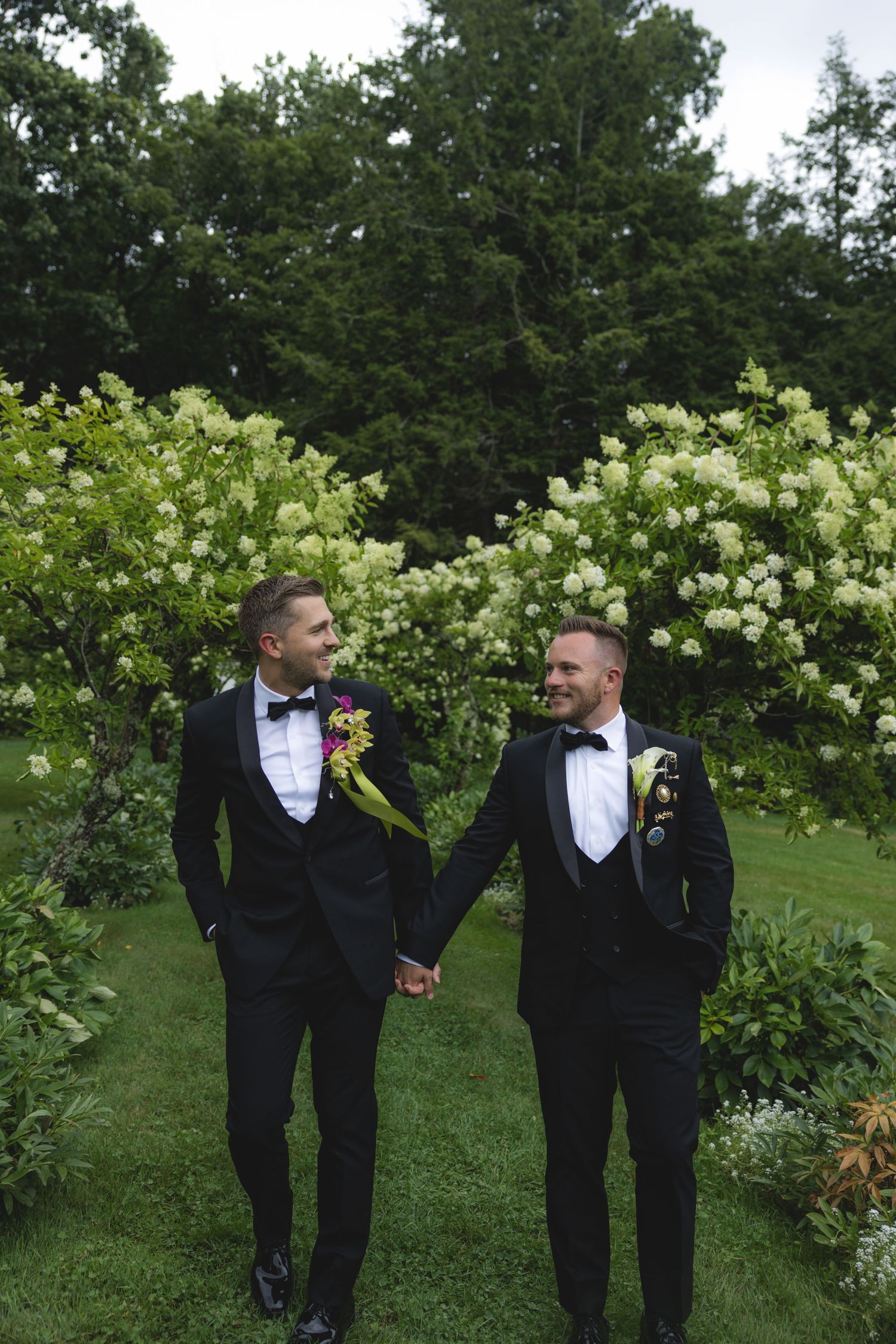 Two grooms in black tuxedos hold hands and smile at each other in a lush garden with white flowering shrubs.