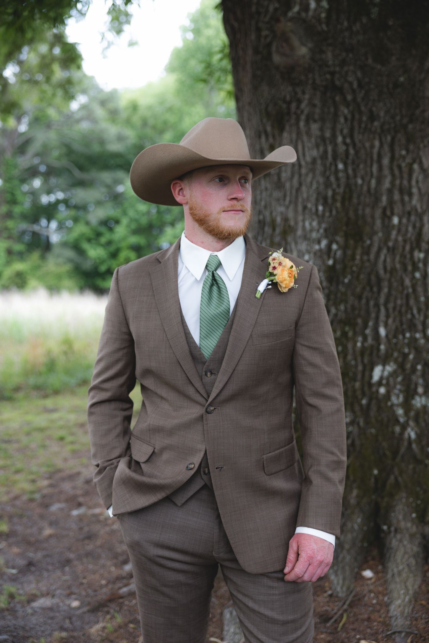 A groom in a brown three-piece suit, green tie, boutonniere, and tan cowboy hat stands outdoors near a tree.