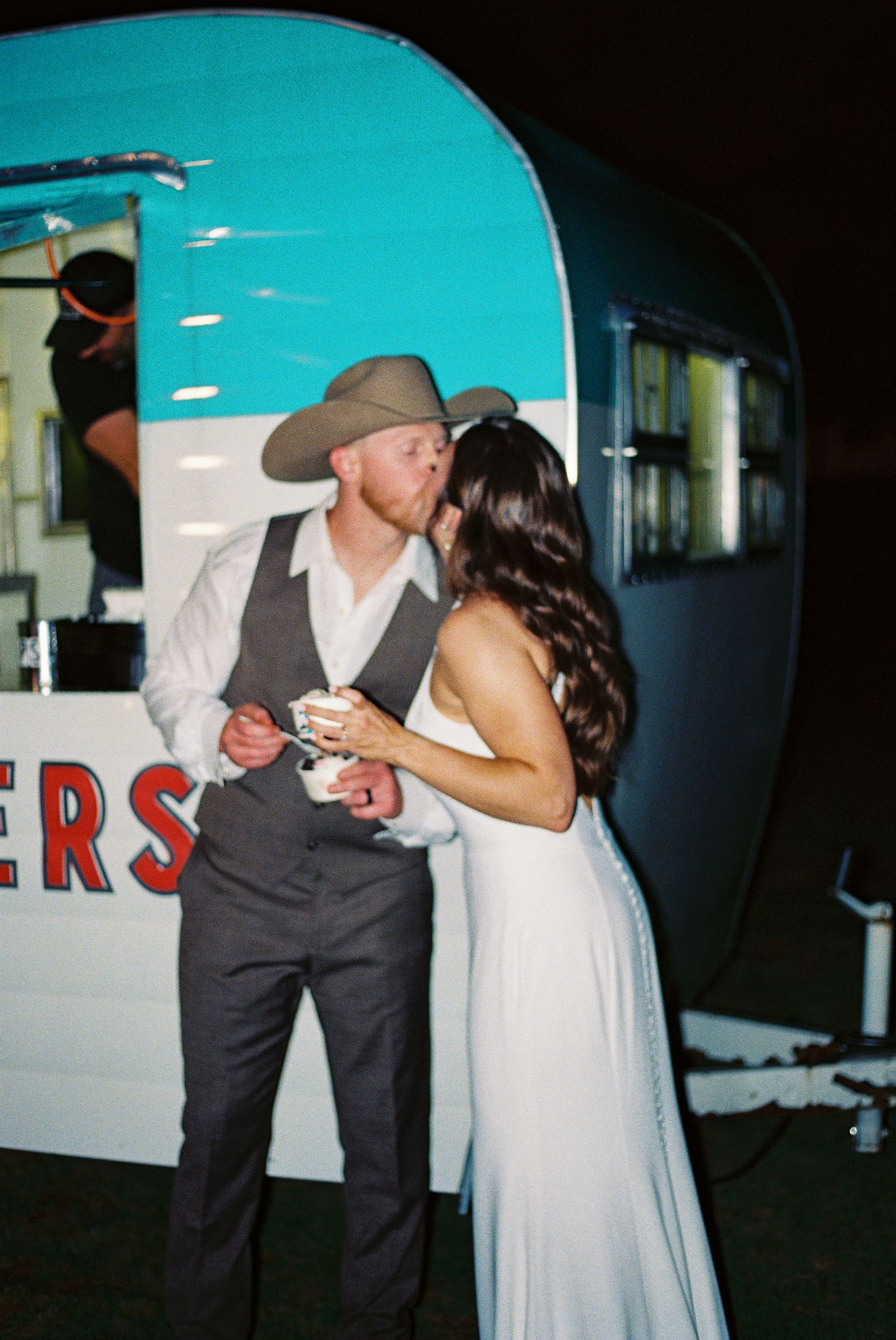 Bride and groom sharing a kiss near a vintage turquoise trailer at their outdoor wedding reception at night.