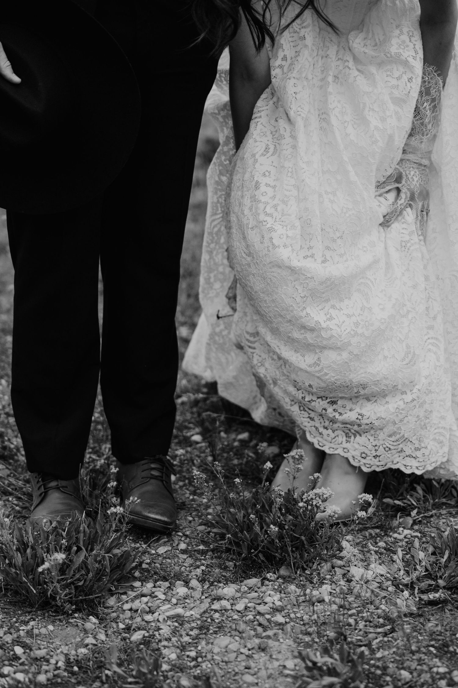 Black and white photo of bride in lace gown and groom in dark suit standing on rocky ground at wedding.