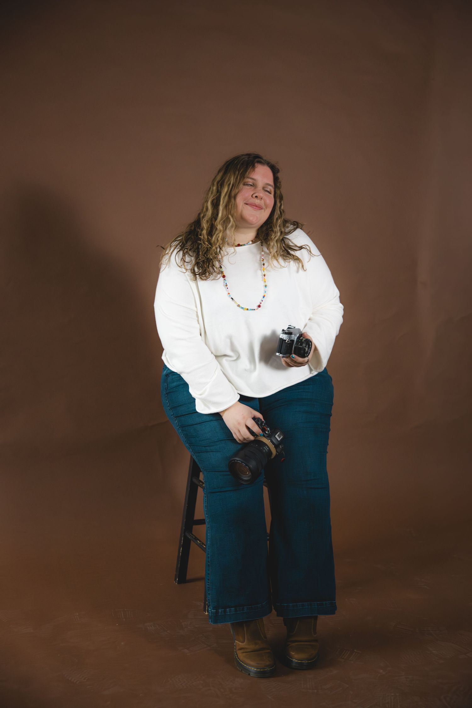 Woman in white sweater and teal wide-leg jeans sits on stool holding cameras in a studio setting.