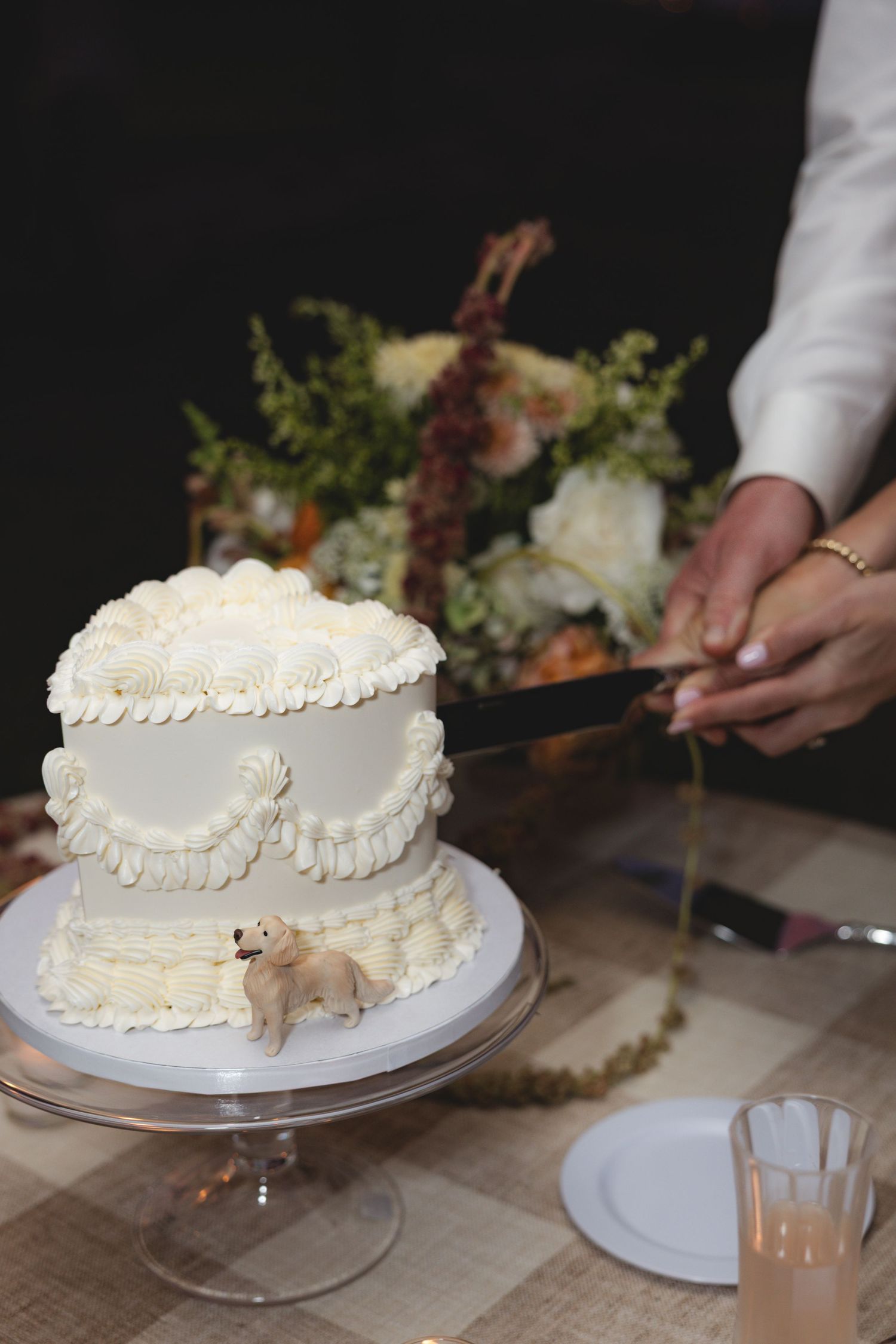 Two-tier white buttercream wedding cake with a golden retriever figurine topper being cut at the reception.