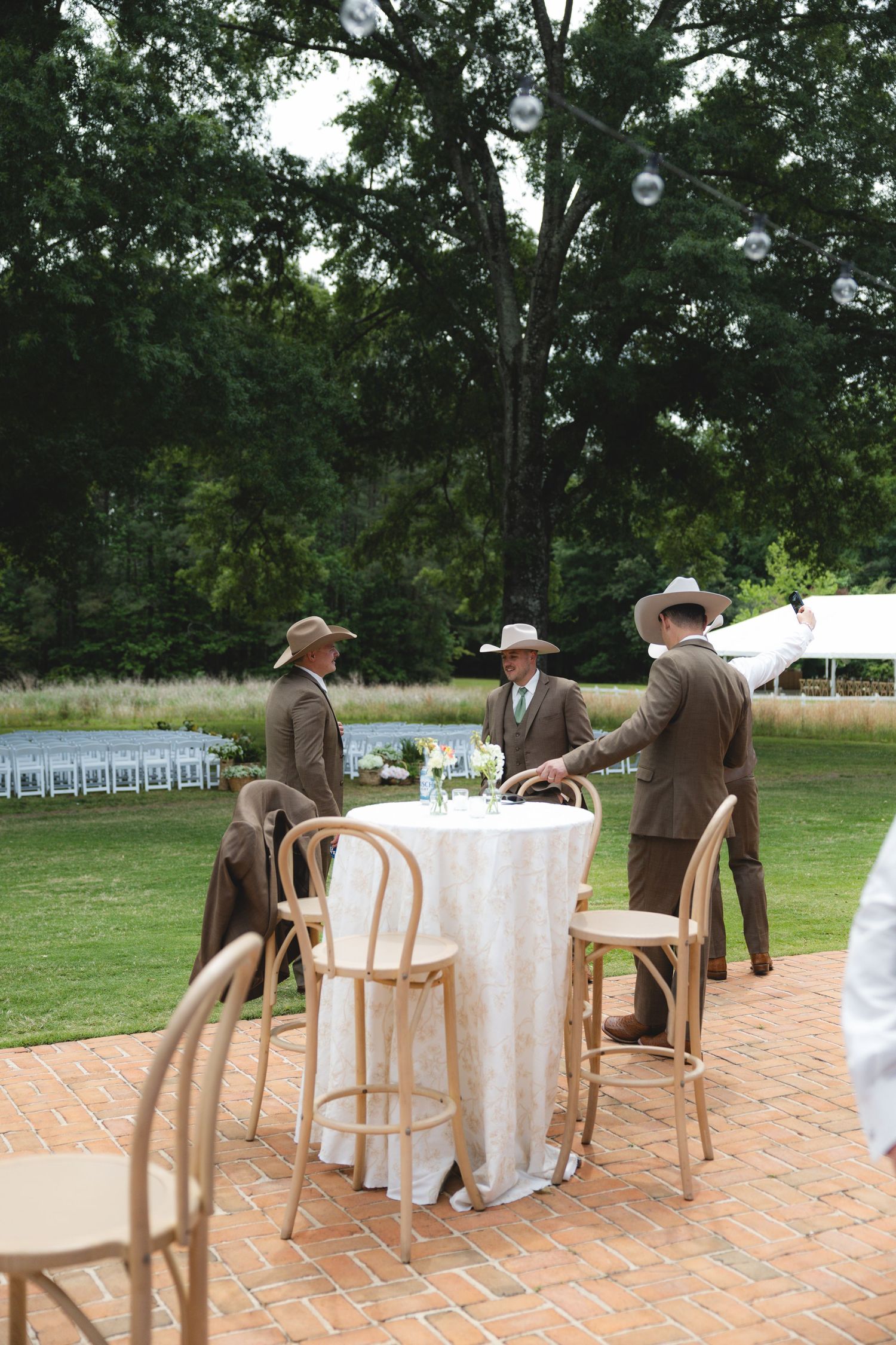 Three groomsmen in brown suits and cowboy hats gathered at cocktail table on brick patio.