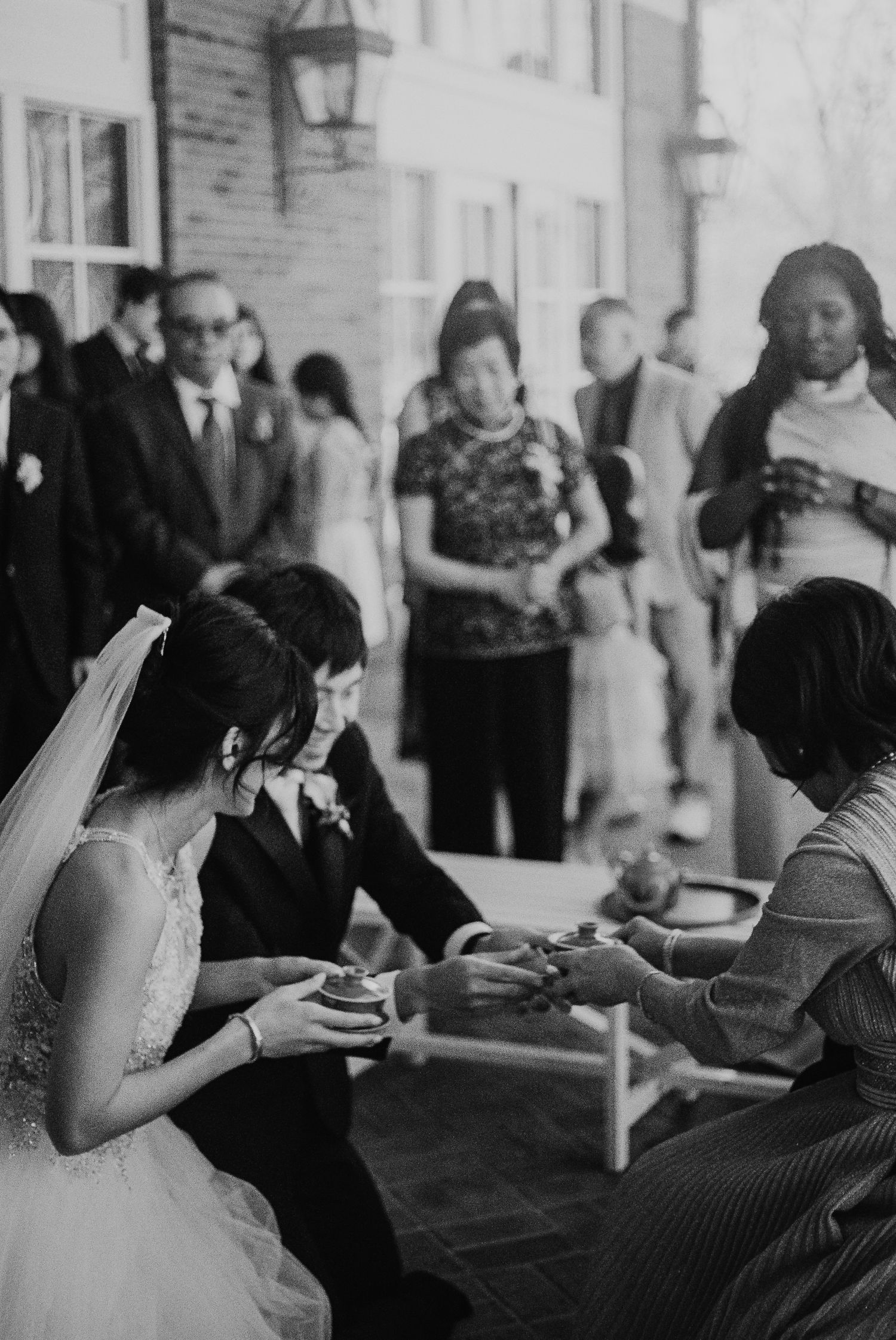 Black and white photo of bride and groom participating in traditional tea ceremony with guests watching.