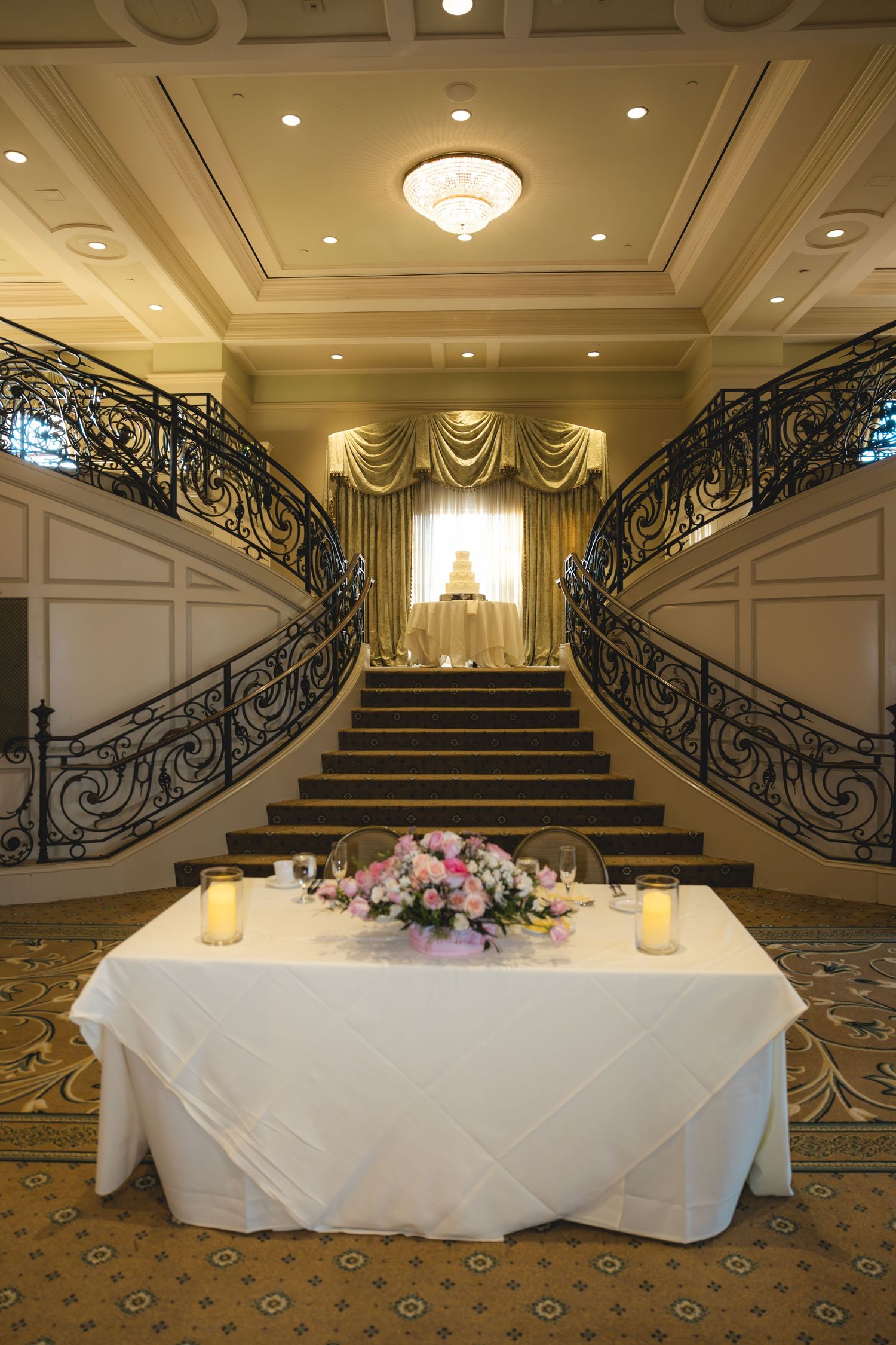 Elegant wedding reception venue with grand dual staircases, sweetheart table, floral centerpiece, and tiered wedding cake backdrop.