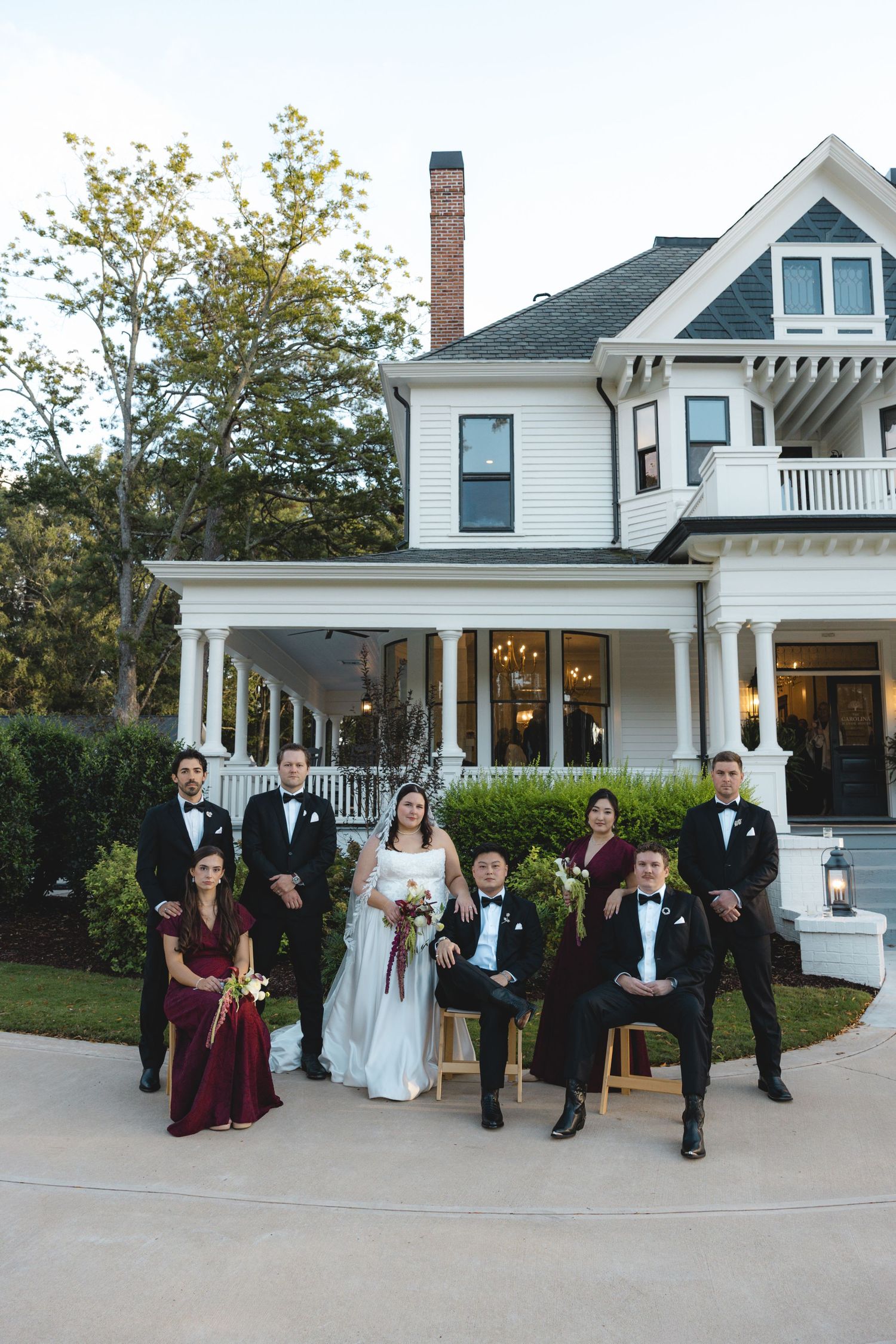 Bride in a white gown poses with groomsmen in black tuxedos and bridesmaids in burgundy dresses in front of a white historic house.