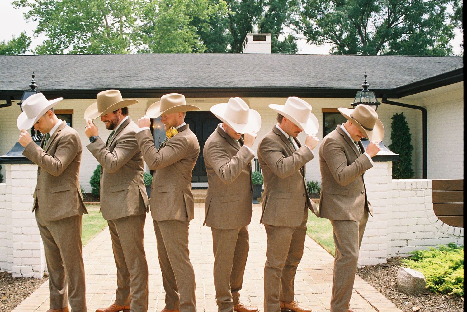 Six groomsmen in tan suits and white cowboy hats adjusting hats outside wedding venue.