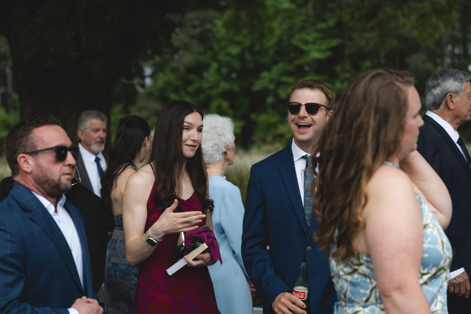 Wedding guests socialize outdoors, with woman in burgundy dress and man in navy suit.