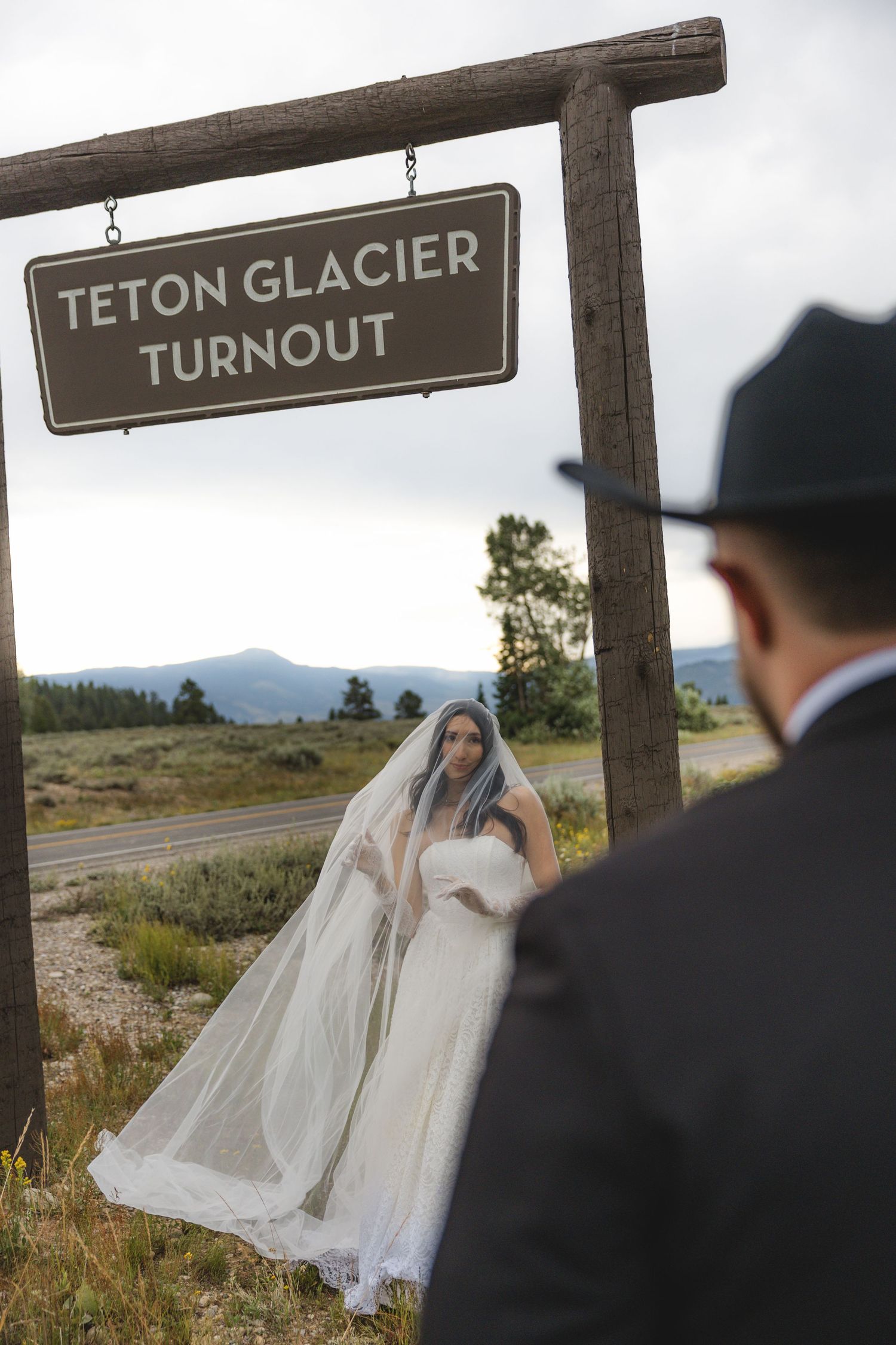 Bride in white gown and veil poses under Teton Glacier Turnout sign as groom watches in Grand Teton National Park.