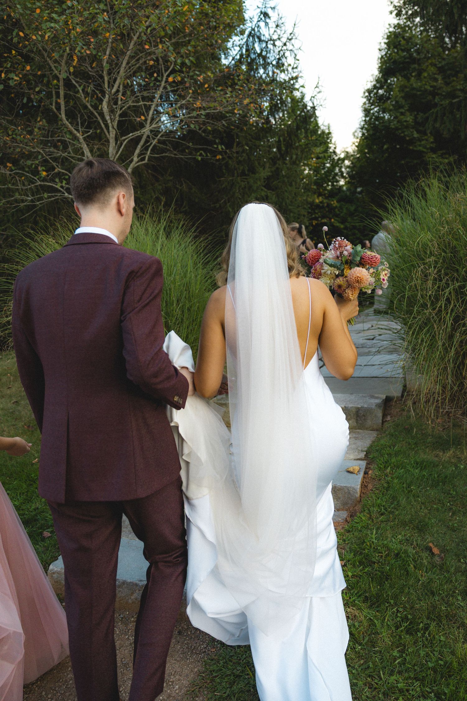 Bride and groom walking hand in hand through a lush garden, bride wearing white gown with veil holding floral bouquet.