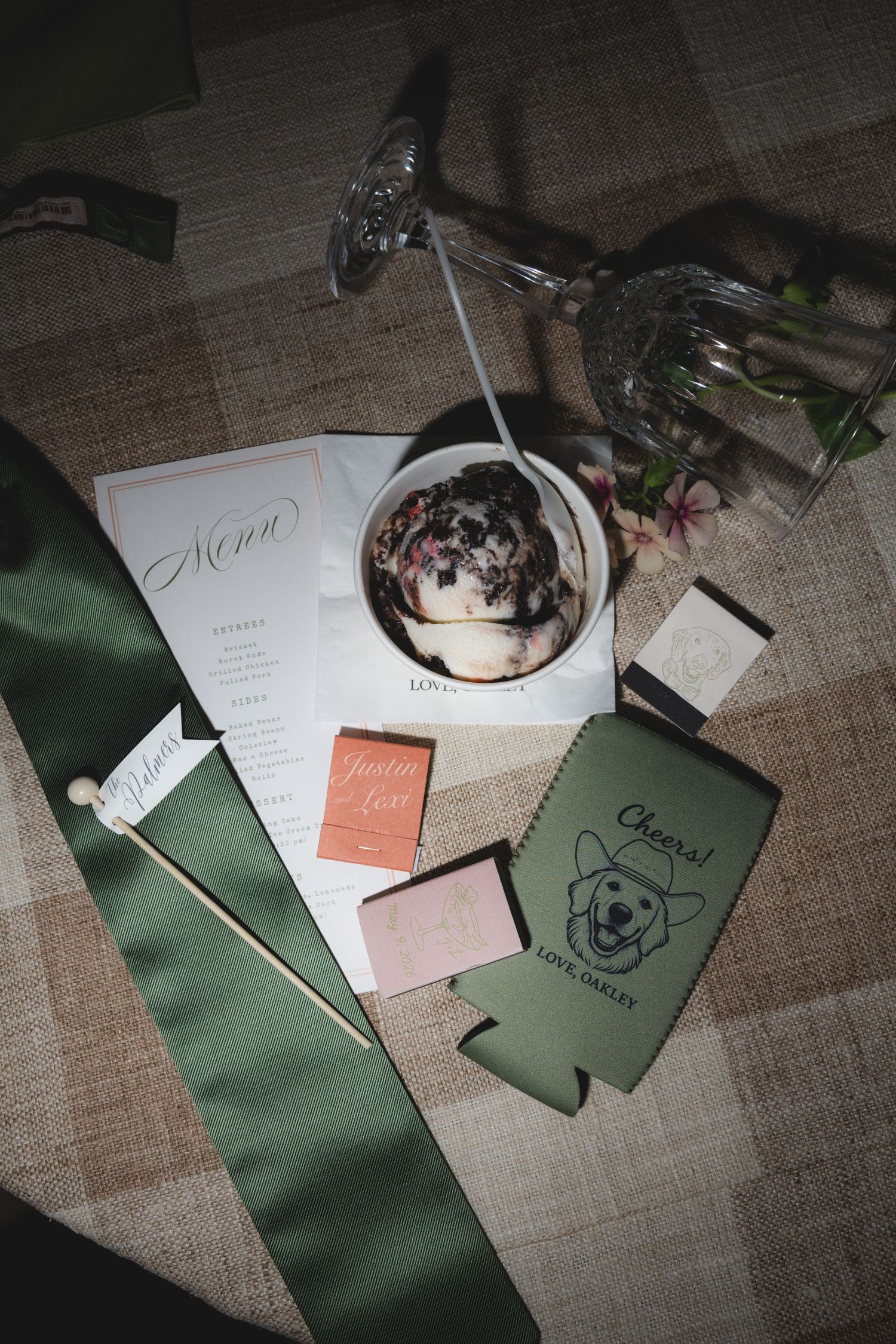 Wedding flat lay with menu, green satin ribbon, koozie, matchbook, ice cream bowl, and crystal glassware on linen.