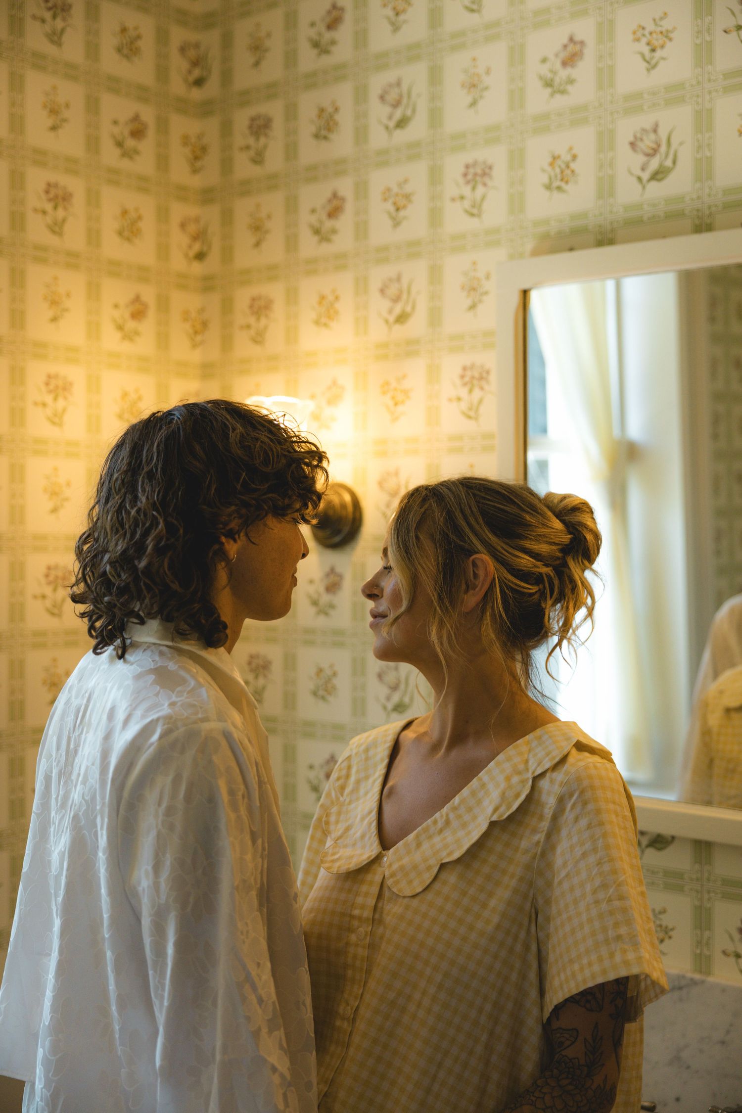 Two young people in vintage pajamas face each other intimately in a warmly lit room with floral wallpaper.