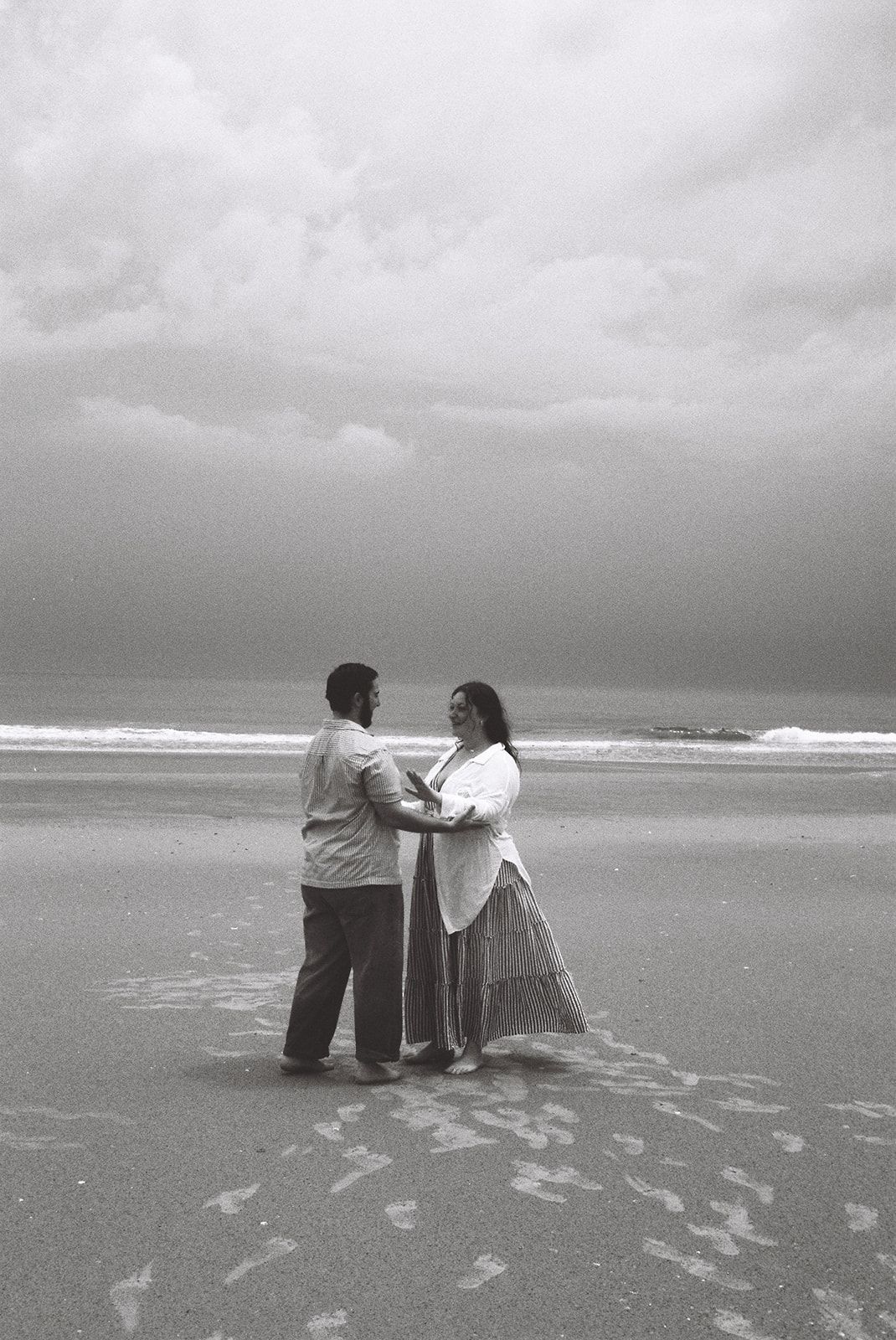 Black and white photo of a couple holding hands and facing each other on a sandy beach under dramatic cloudy skies.