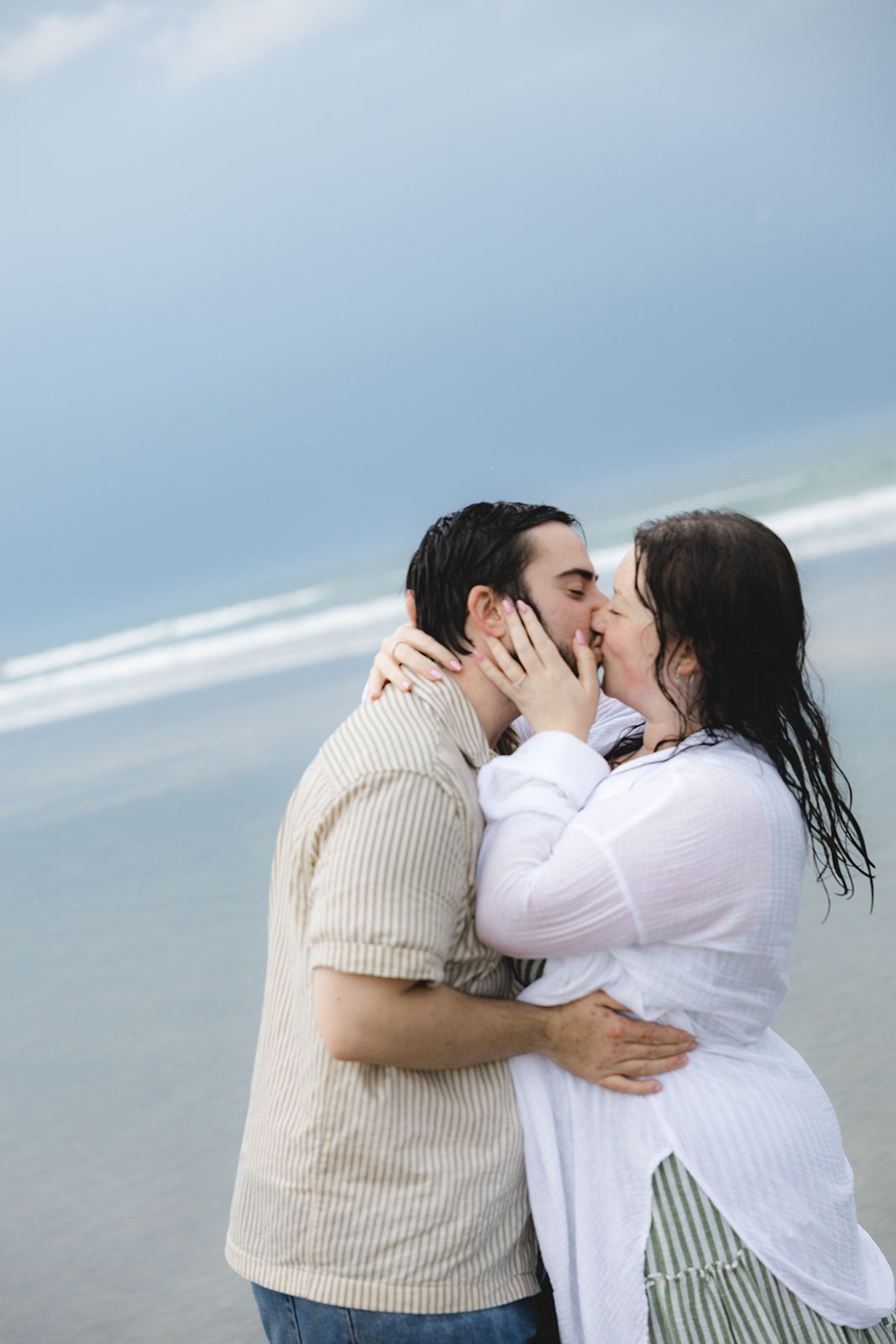 Romantic couple sharing a passionate kiss on a beach, with ocean waves and cloudy blue sky in the background.