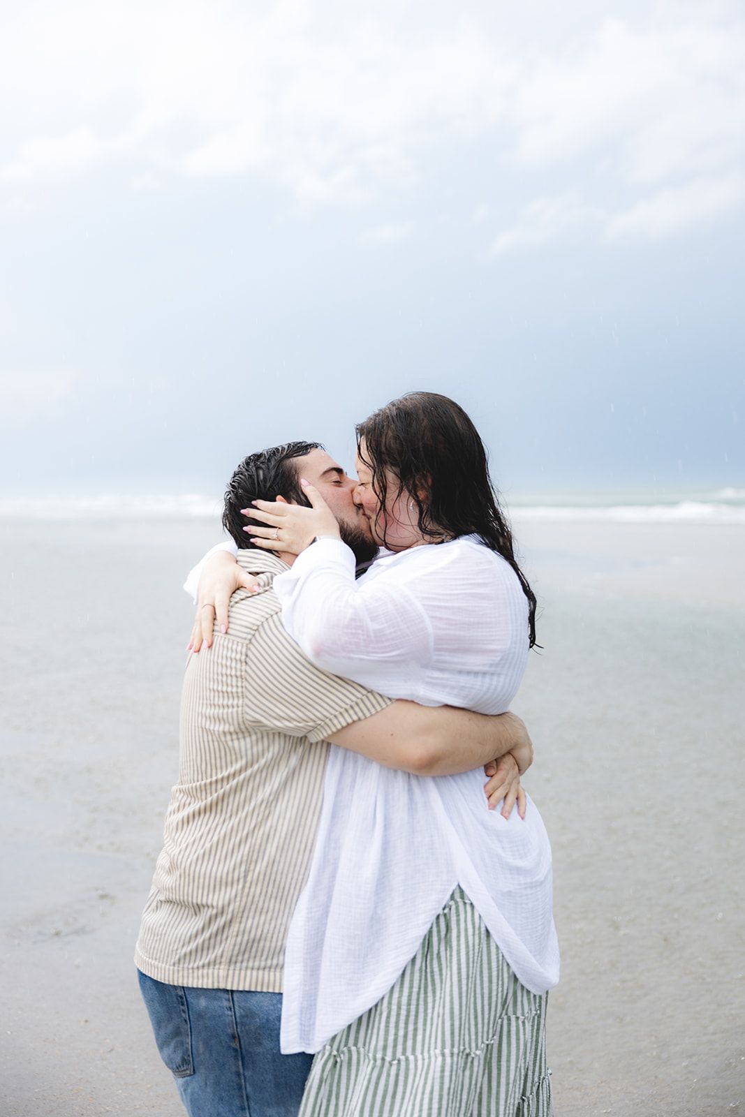 Couple sharing a romantic kiss on a cloudy beach, embracing each other in casual summer attire.