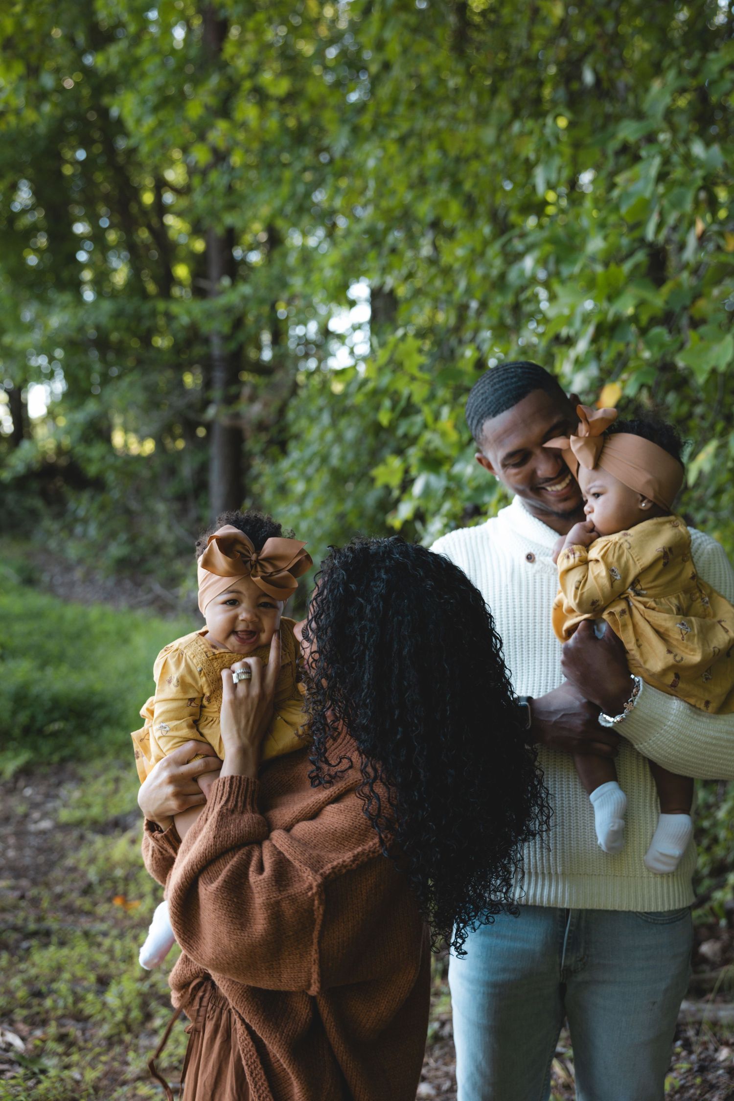 A happy family of four enjoys an outdoor autumn stroll, with parents holding two young children dressed in warm yellow outfits.
