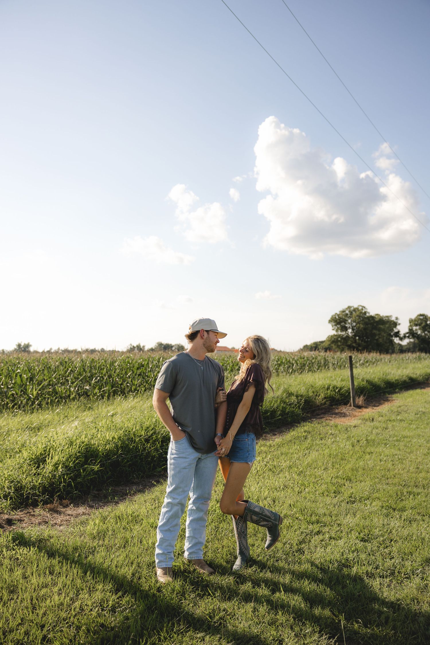 A couple holds hands on a rural path beside a cornfield under a partly cloudy sky on a sunny day.