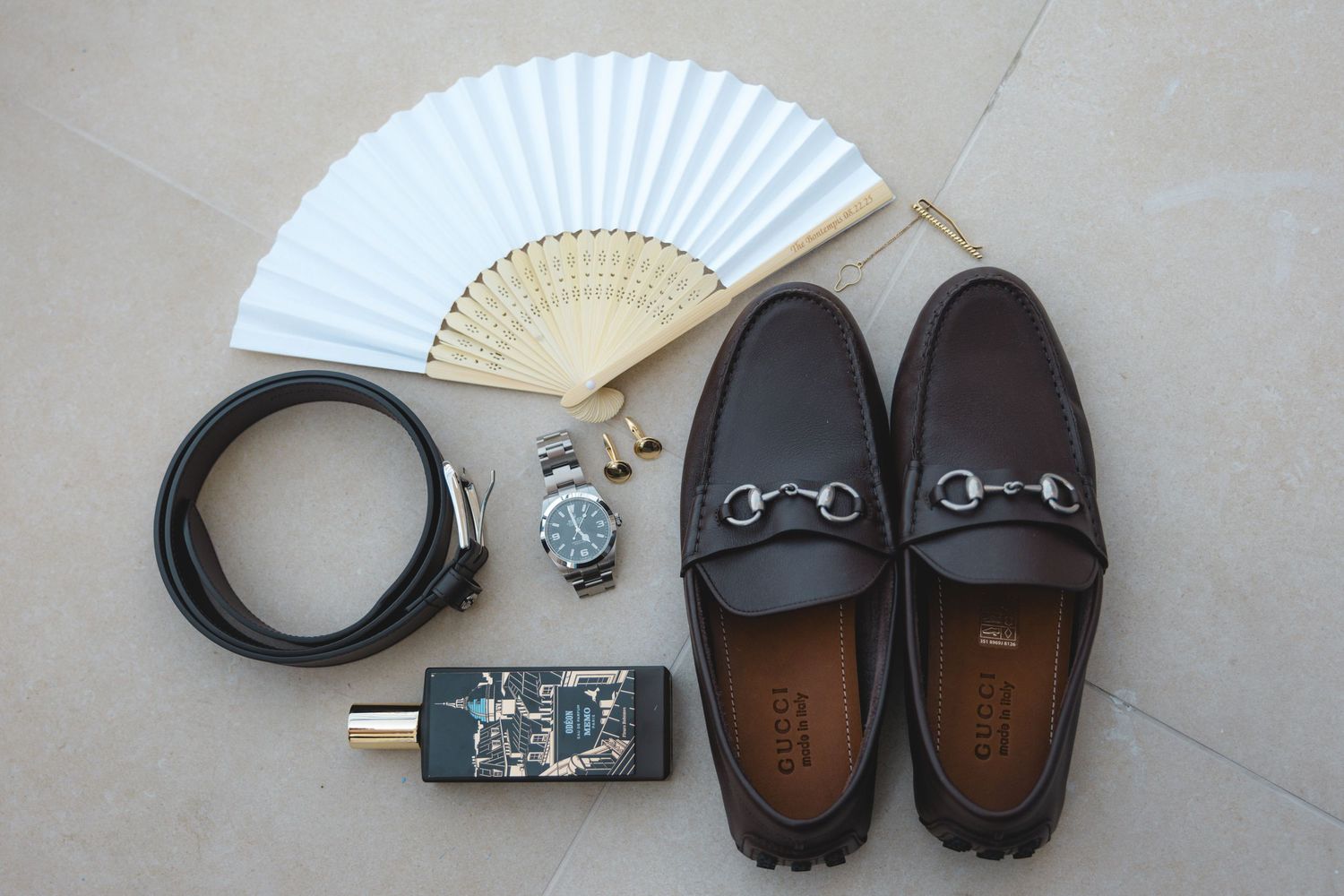 Flat lay of men's accessories including brown loafers, black belt, watch, cologne, and white fan on gray surface.