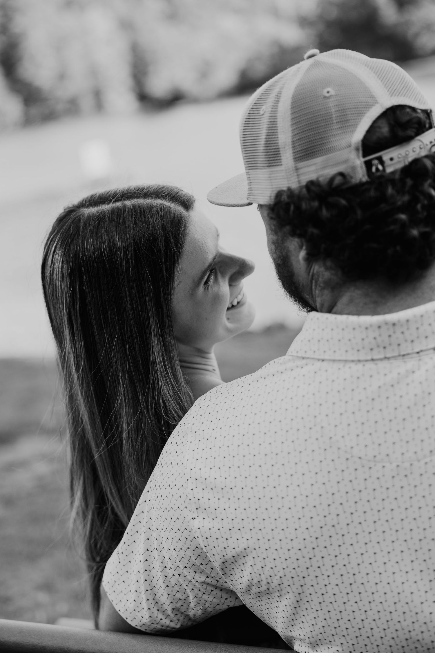 Black and white photo of a smiling woman gazing up at a man wearing a trucker hat, sitting together outdoors.