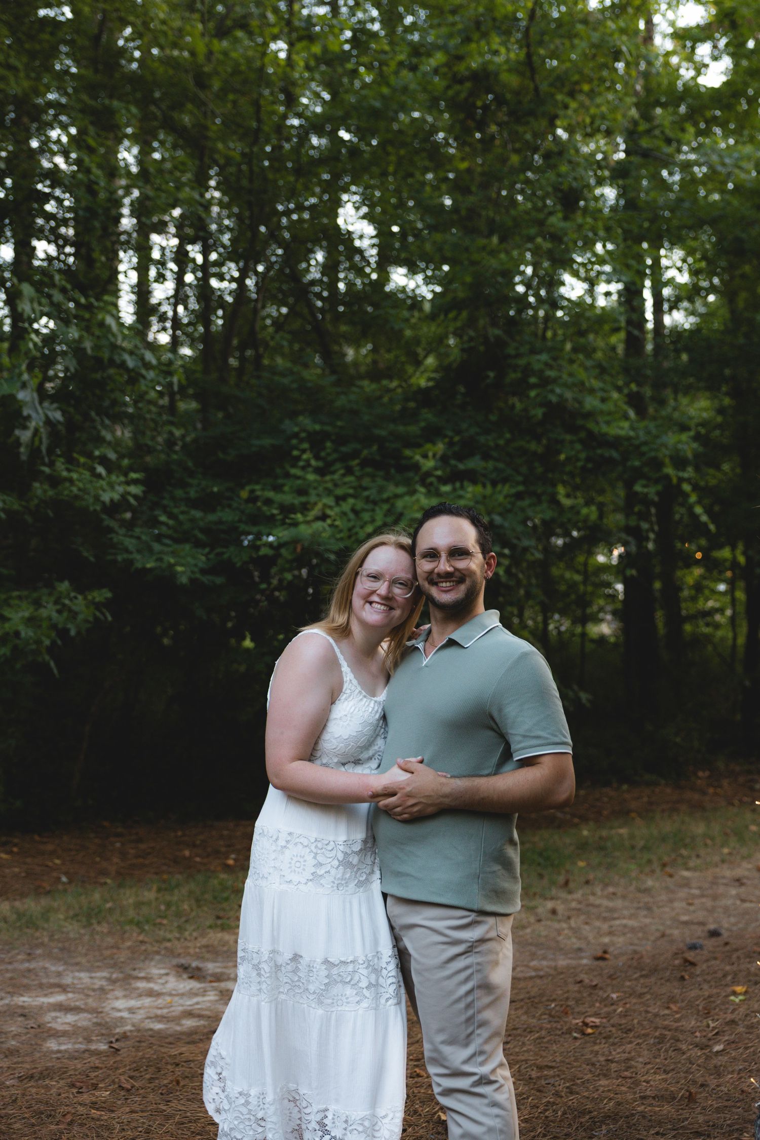 A smiling couple embraces outdoors in a wooded setting, woman in white lace dress, man in sage polo shirt.