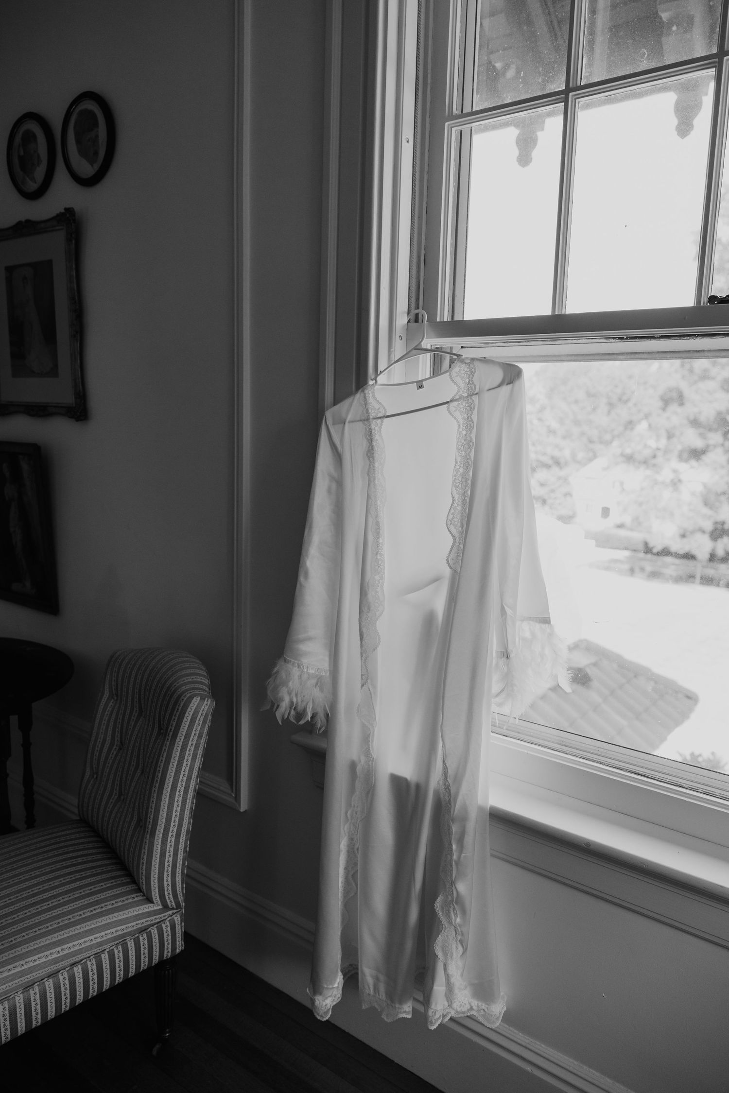 White lace bridal robe hanging on a window hanger in a vintage room with striped chair, black and white photo.