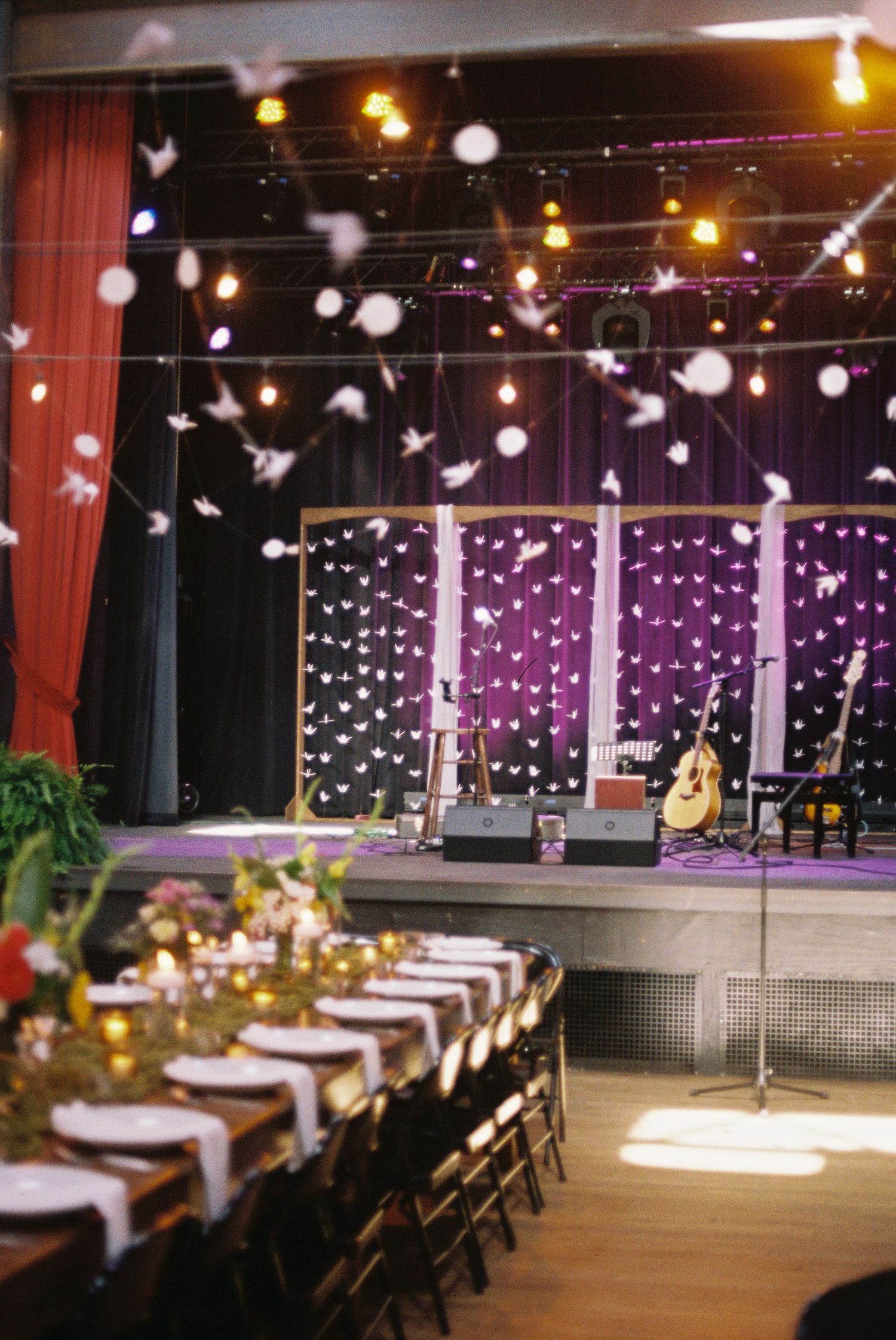 Wedding reception venue with stage, string lights, hanging paper cranes, and elegantly set dining tables.