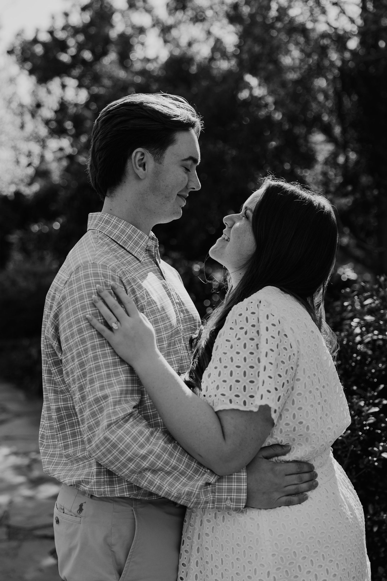 Black and white photo of a young couple embracing and smiling at each other outdoors among lush greenery.