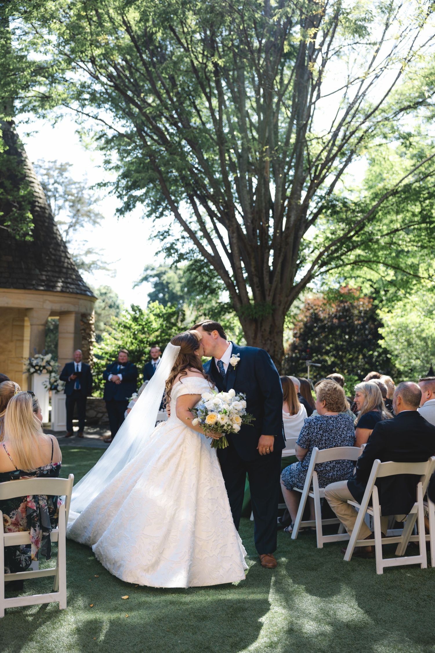 Bride and groom share first kiss as newlyweds during outdoor garden wedding ceremony with guests seated on white chairs.