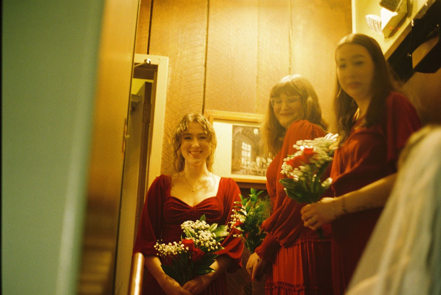 Three women in formal attire stand together holding bouquets of flowers in a doorway.