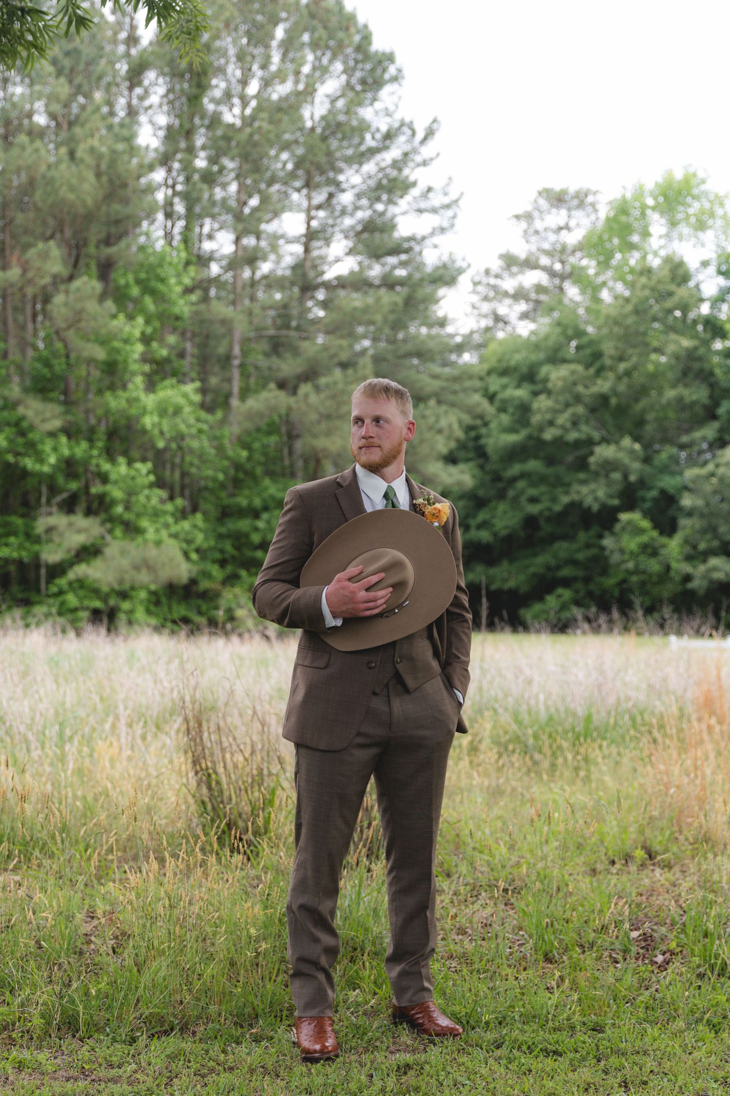 Groom in brown three-piece suit holds tan cowboy hat while standing in a grassy field with trees behind.