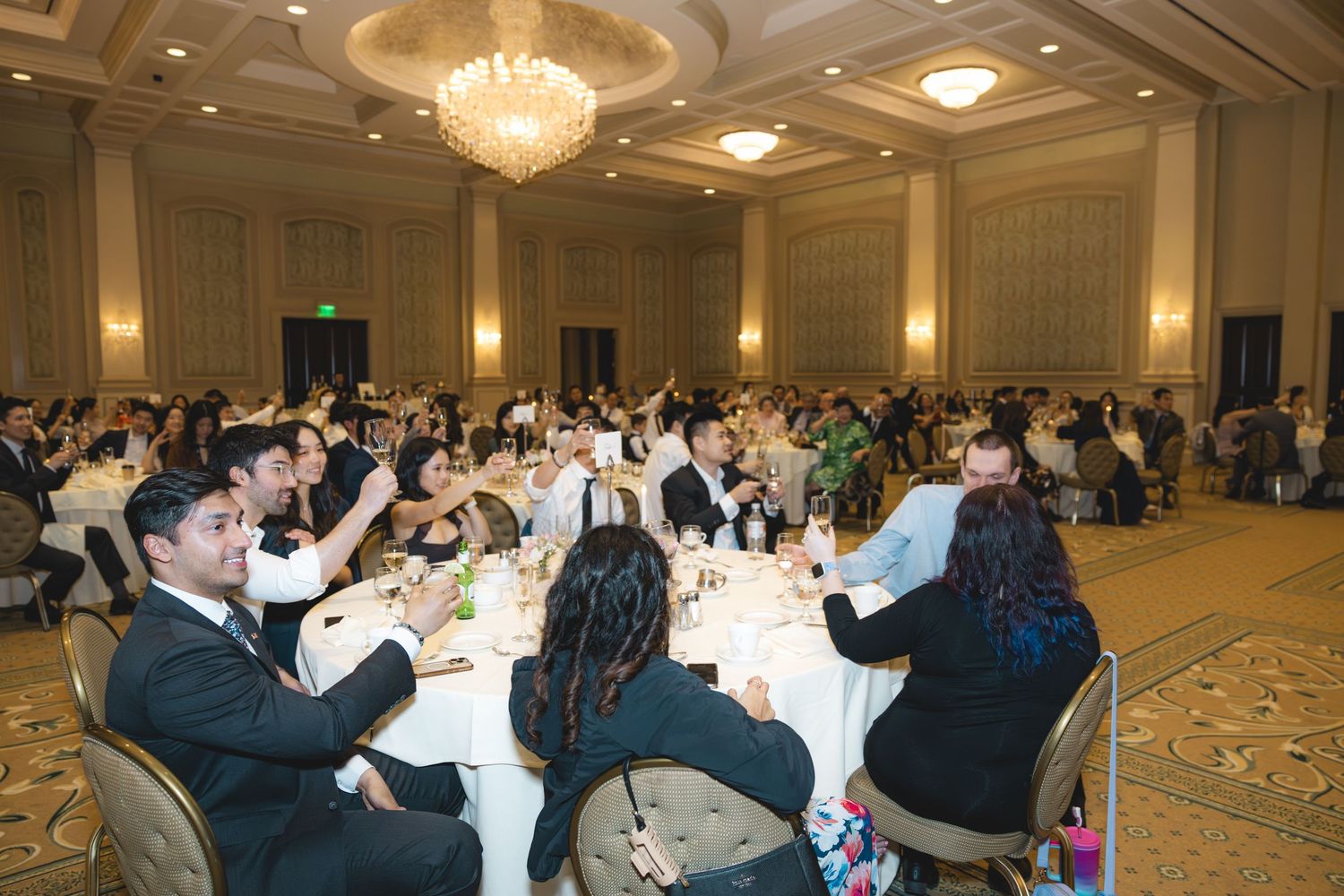 Wedding guests raising glasses for a toast in a grand chandelier-lit ballroom.