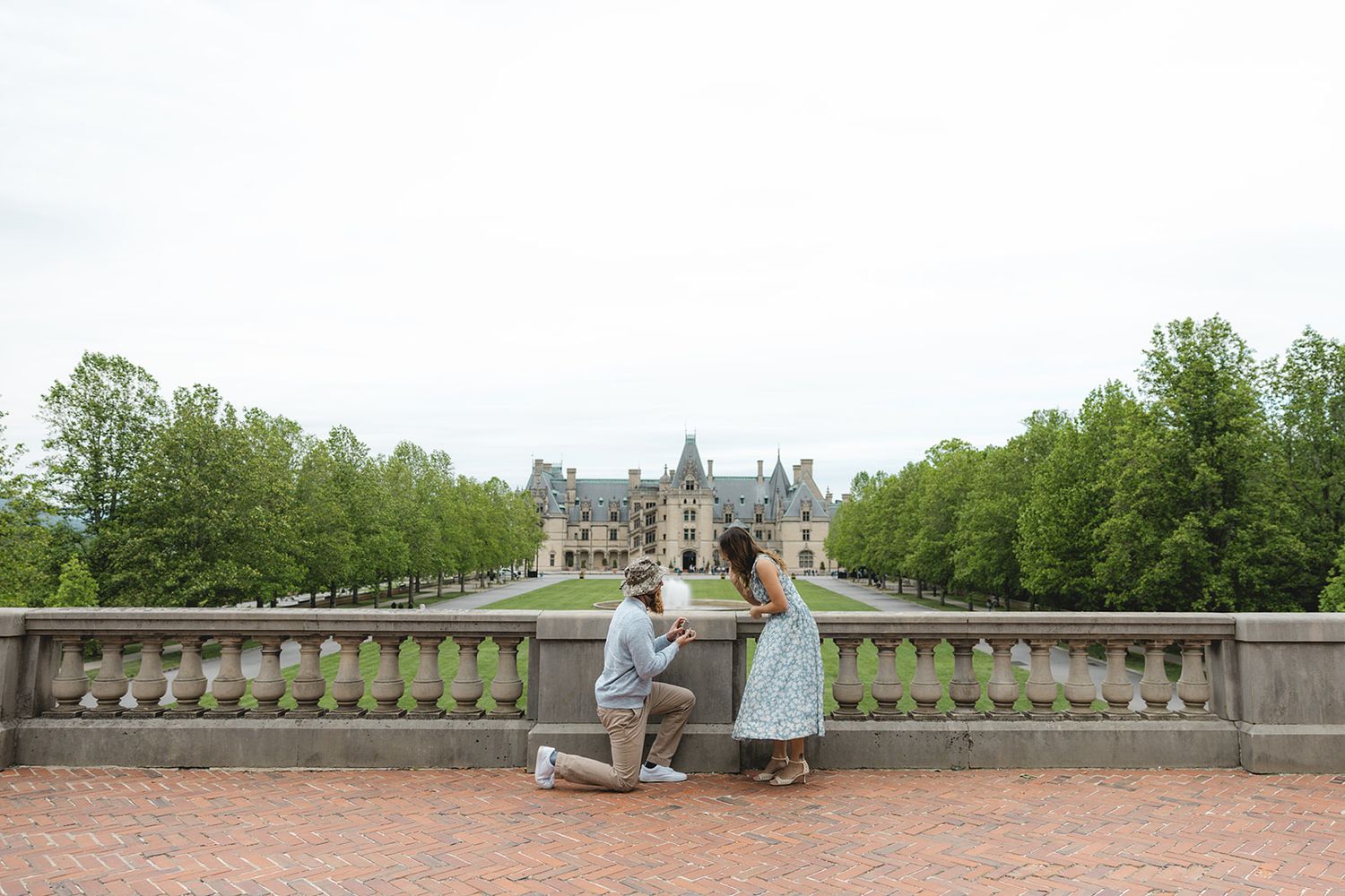 Man proposing to woman on brick terrace in front of grand Biltmore Estate castle surrounded by lush green trees.