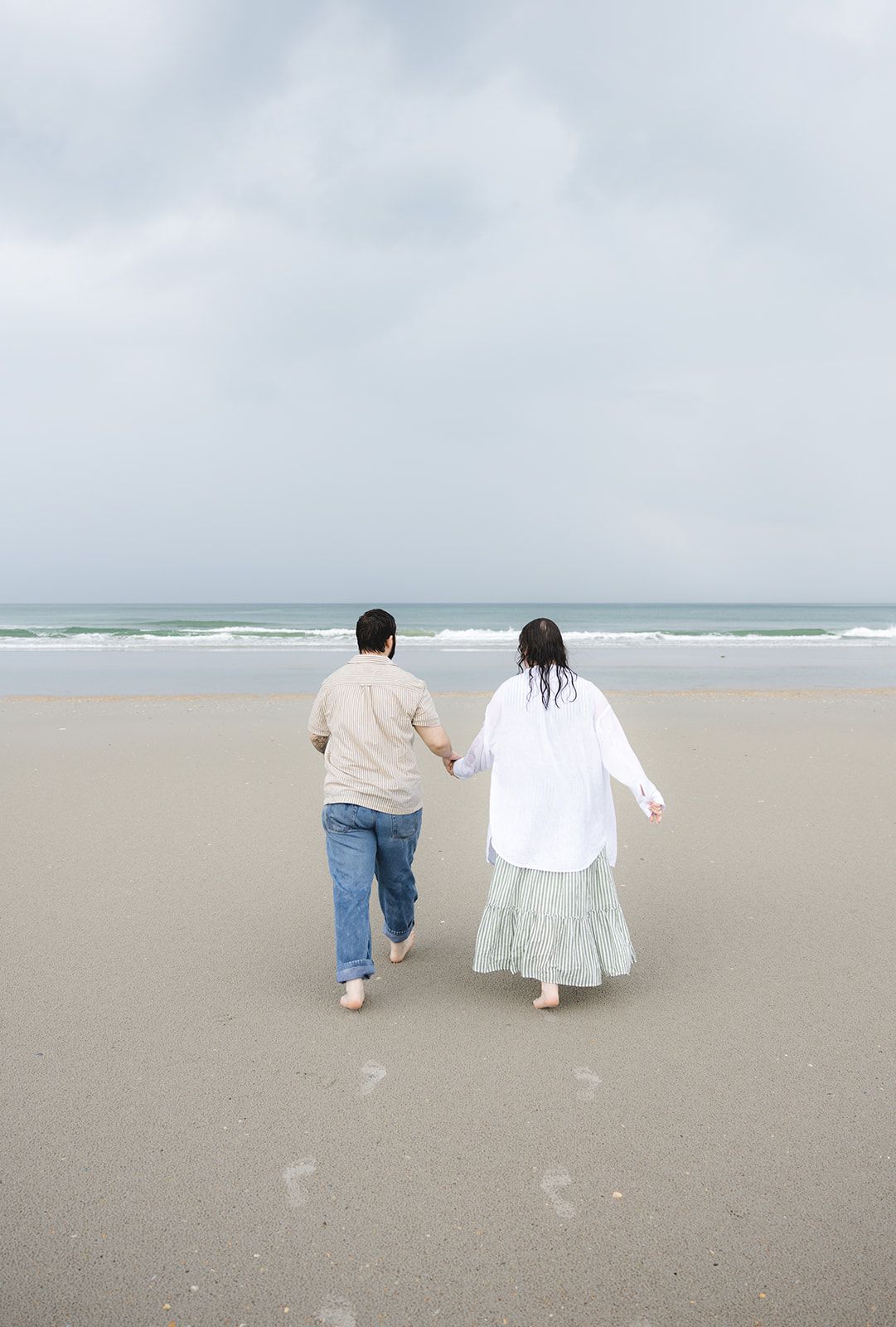 Couple holding hands walking barefoot on a sandy beach toward ocean waves under a cloudy sky.