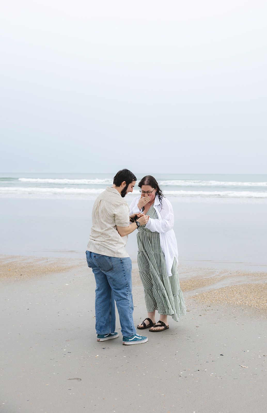 Couple sharing a romantic moment on a sandy beach, woman in green dress and white shirt, man in jeans, ocean waves behind.